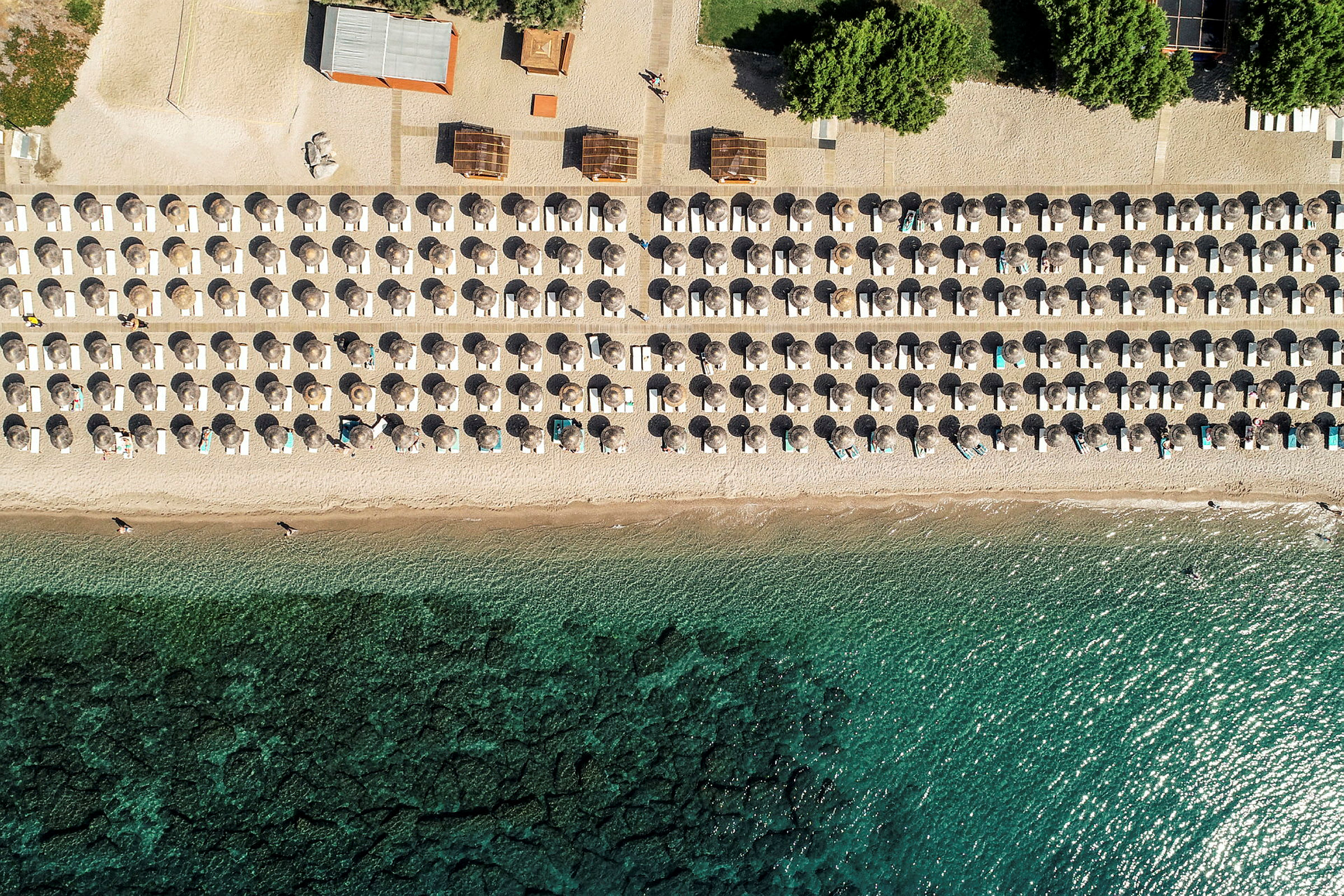 Luftfoto af en strand med parasoller og liggestole placeret i symmetriske rækker ved stranden. (AI-genereret tekst)