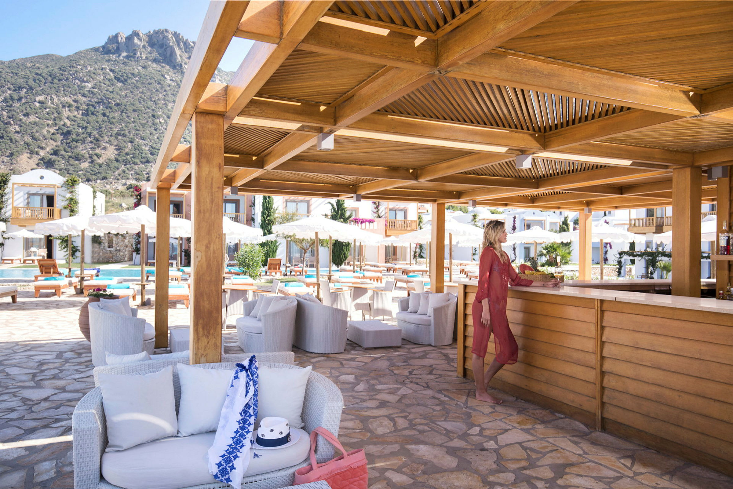 Poolområde med bar og siddepladser under en træpergola. Bjerge i baggrunden. (AI-genereret tekst)