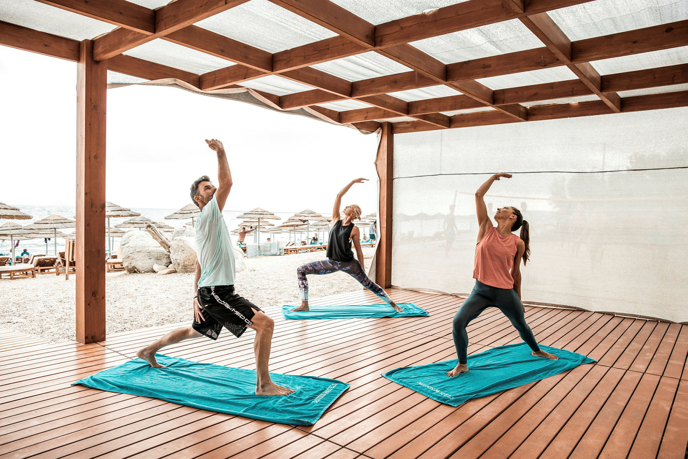 Tre personer udfører yogaøvelser under en trækonstruktion nær en strand. (AI-genereret tekst)