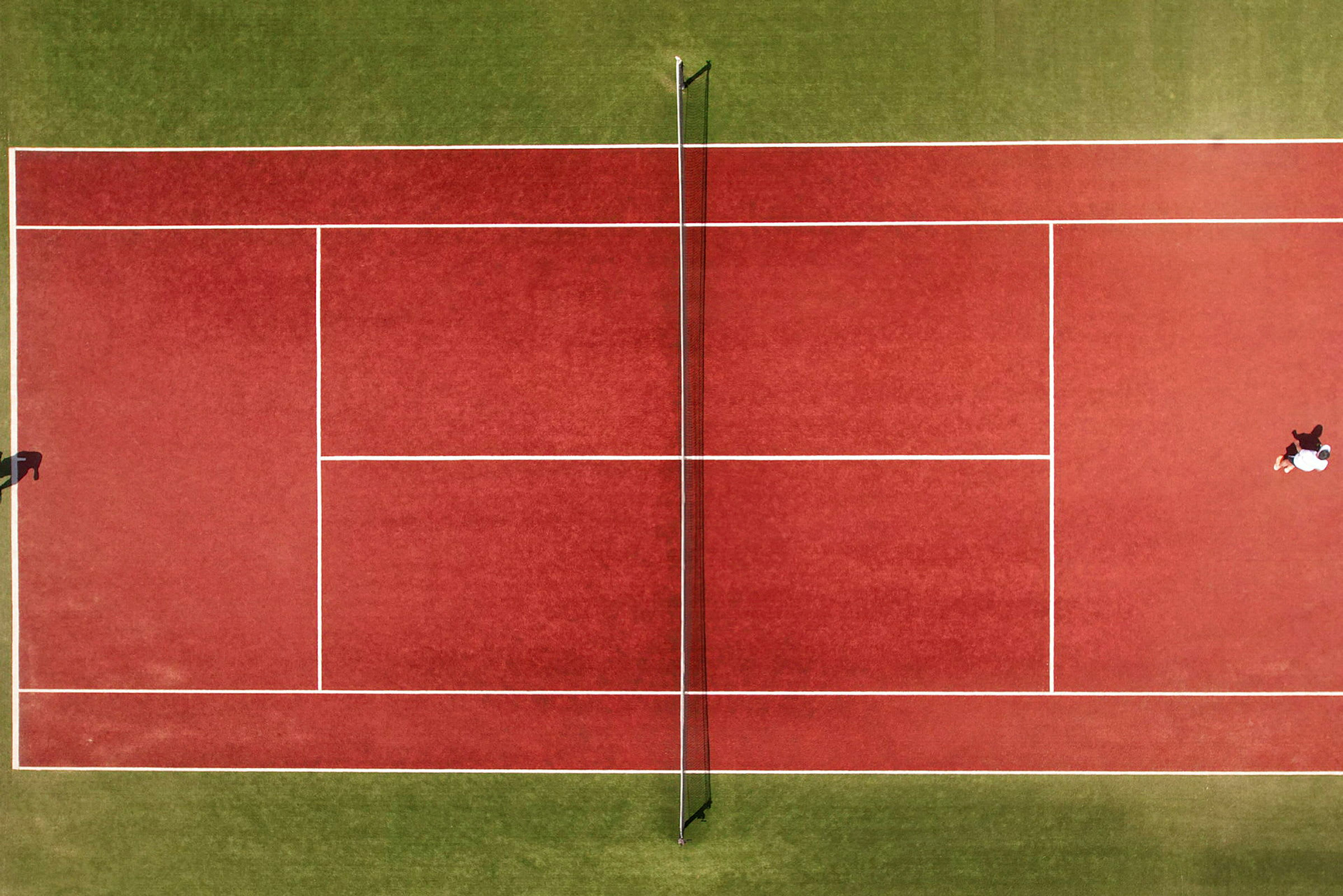 Luftfoto af en rød tennisbane med net, to personer spiller. (AI-genereret tekst)