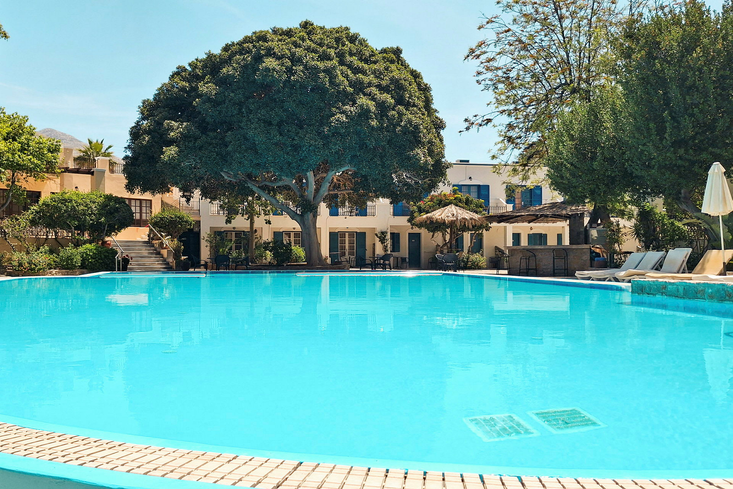 En stor swimmingpool foran et hotel omgivet af grønt og parasoller. (AI-genereret tekst)