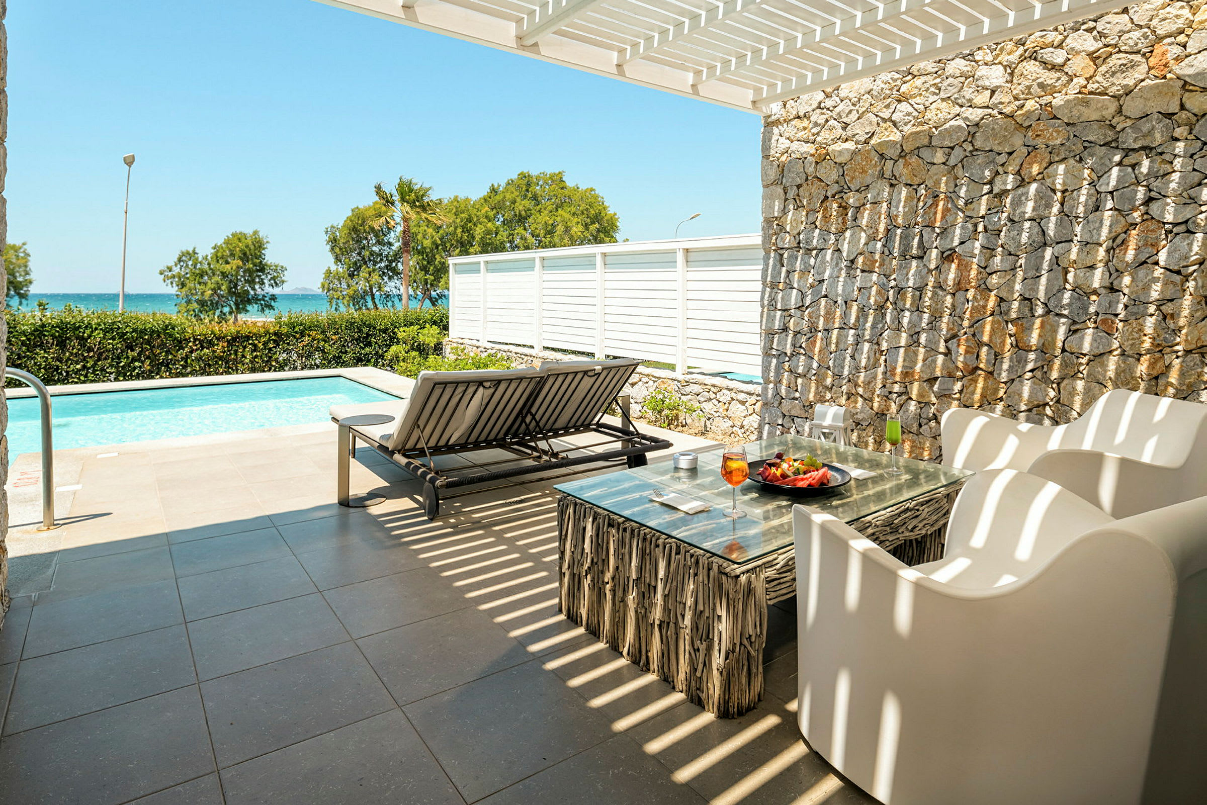 Terrasse med havemøbler og solseng ved en pool, med havudsigt i baggrunden. (AI-genereret tekst)