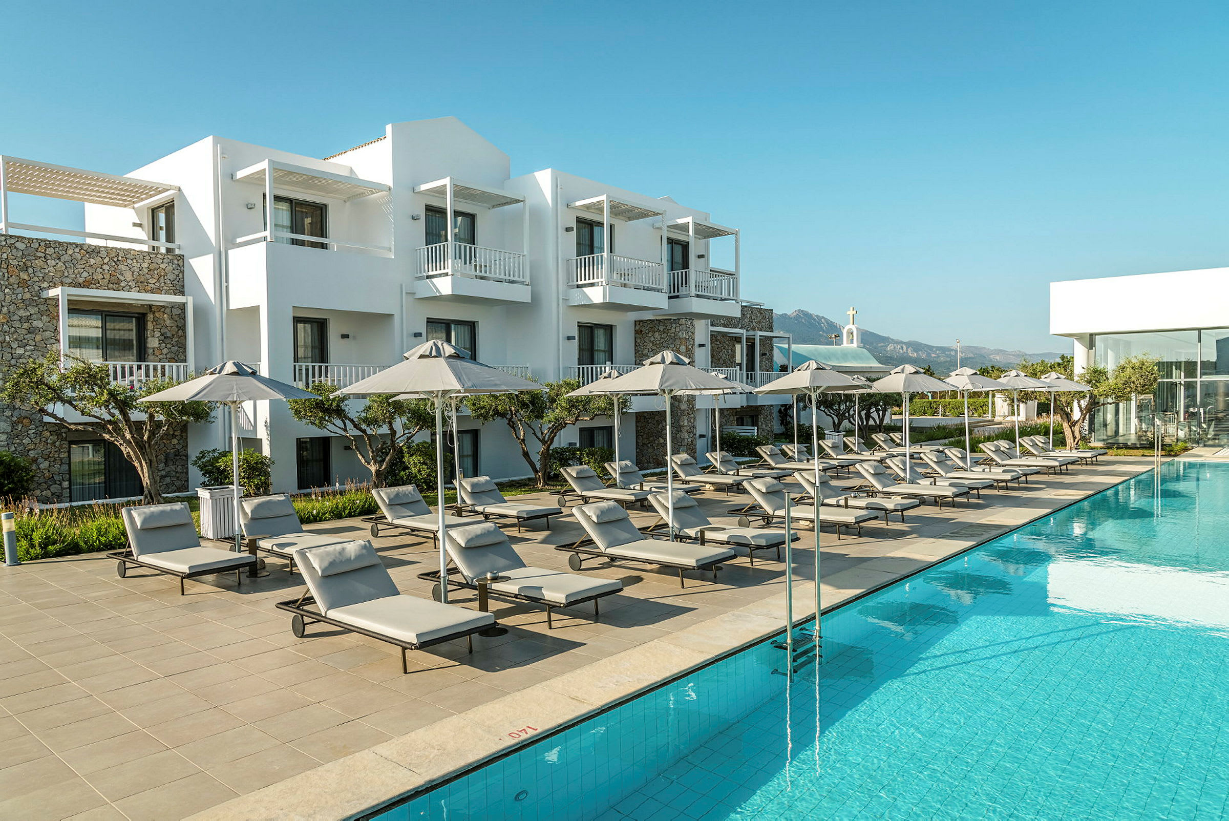 En hotelpool med solsenge og parasoller, omgivet af hvide bygninger. (AI-genereret tekst)