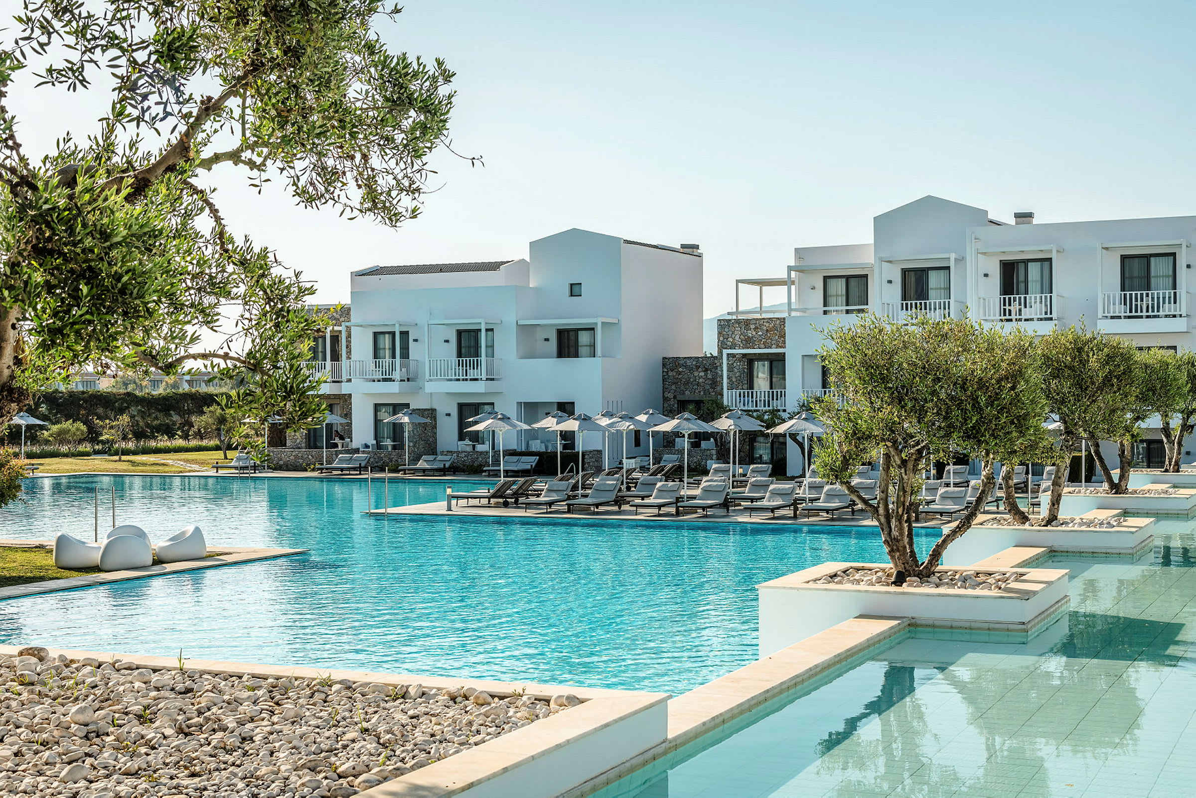 Billedet viser et hotel med hvid facade ved en stor pool, omgivet af solsenge og parasoller samt grønne områder. (AI-genereret tekst)