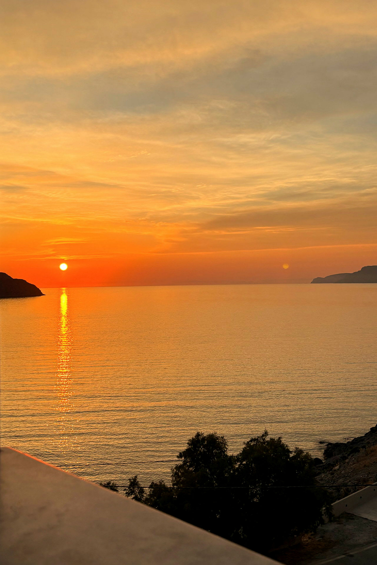 En solnedgang over havet med orange og gule farver på himlen, nogle træer i forgrunden og en klippe til venstre. (AI-genereret tekst)