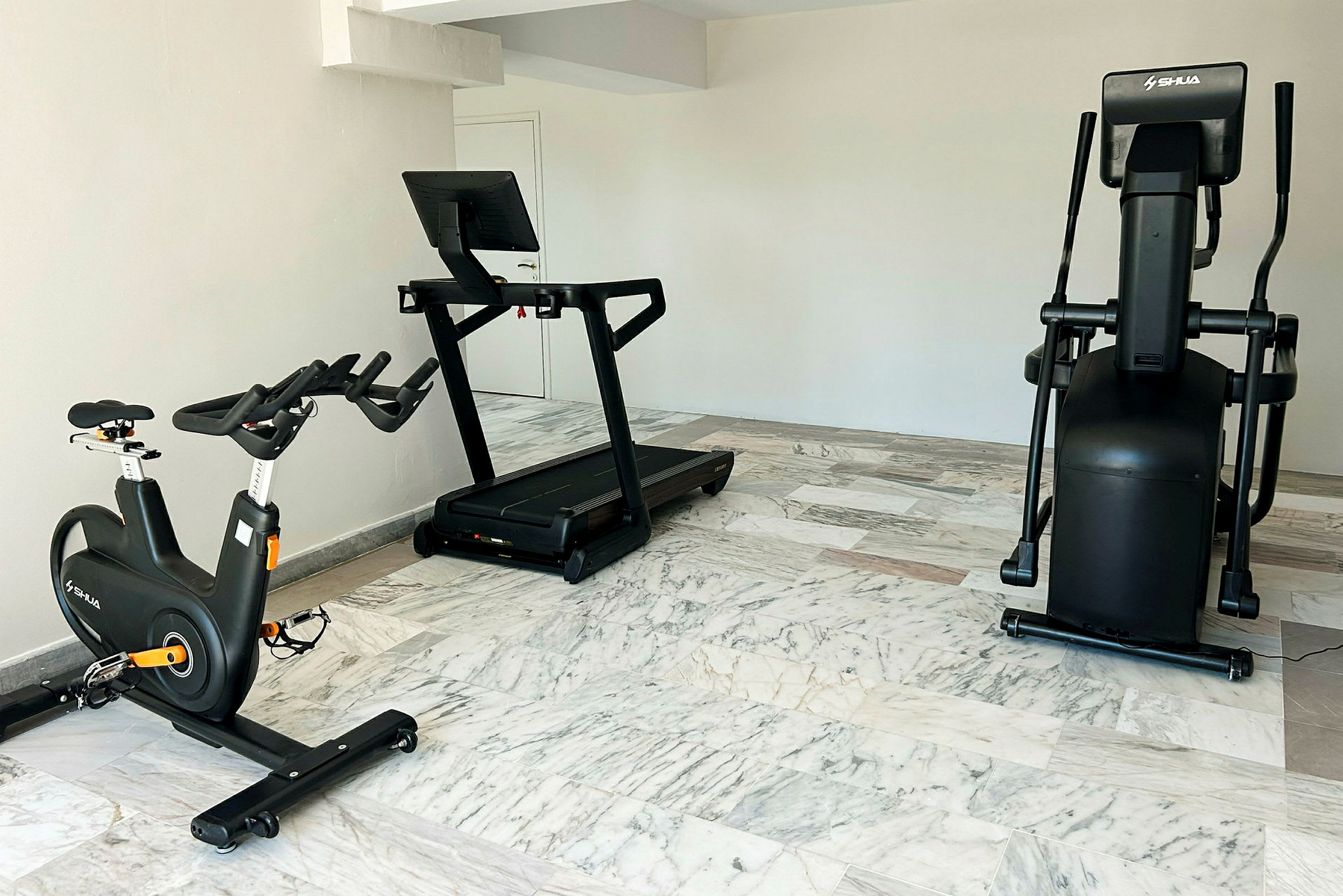 Billede af et fitnessrum med en motionscykel, et løbebånd og en crosstrainer placeret på et gulv med marmorgulvfliser. (AI-genereret tekst)