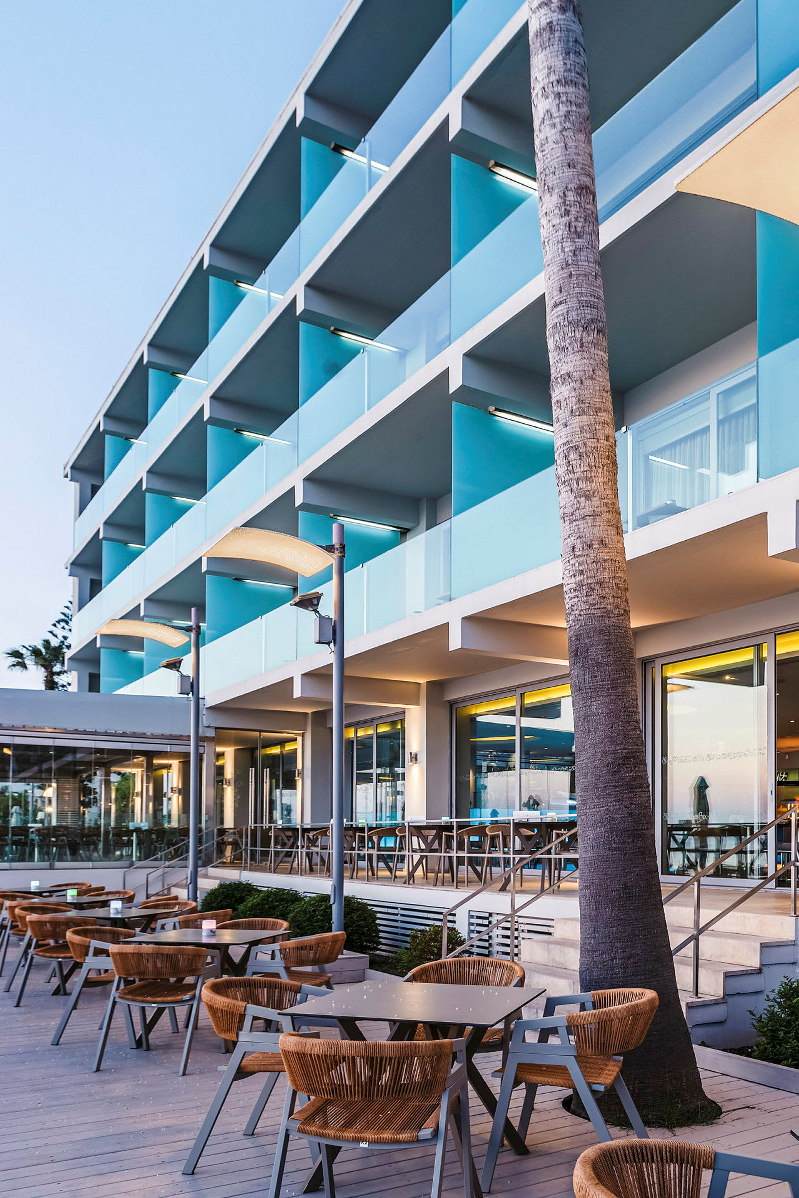 Billede af en moderne hotellfasade med altaner og udendørsservering ved solnedgang. (AI-genereret tekst)