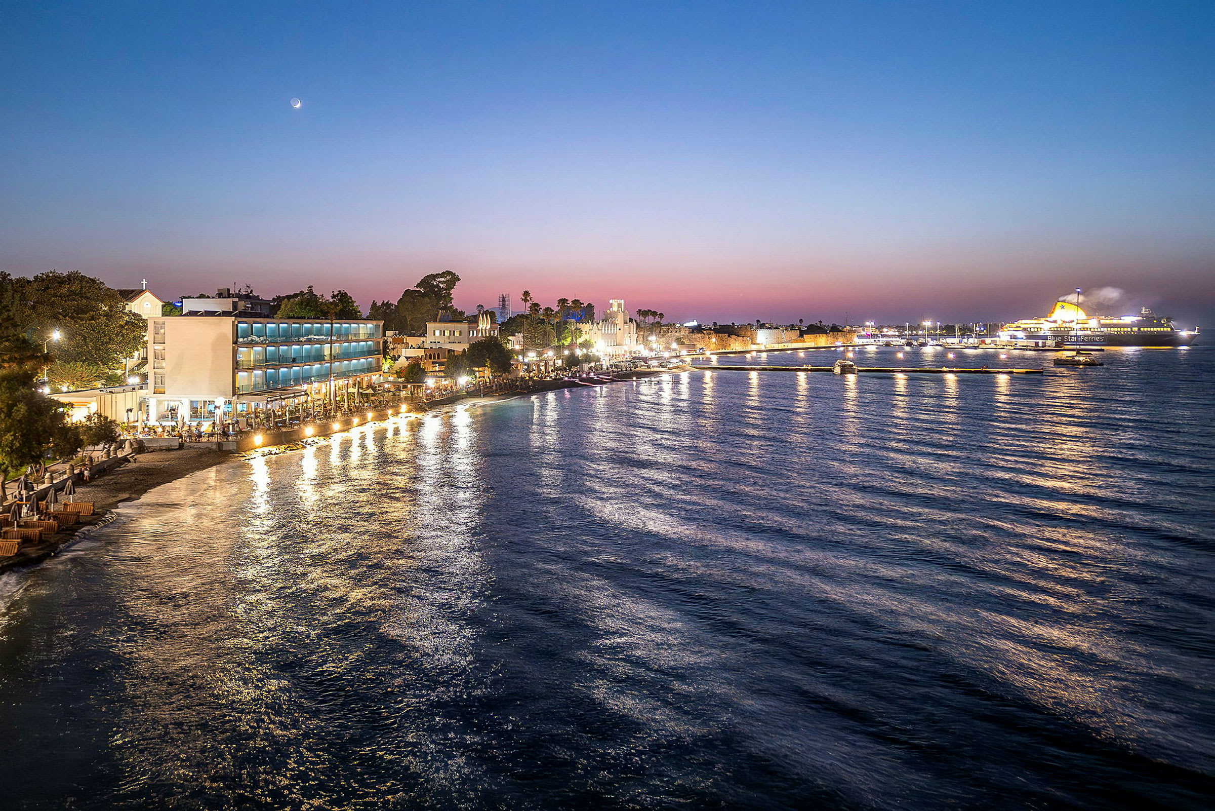 Udsigt over en strandpromenade med bygninger og en færge ved solnedgang. (AI-genereret tekst)