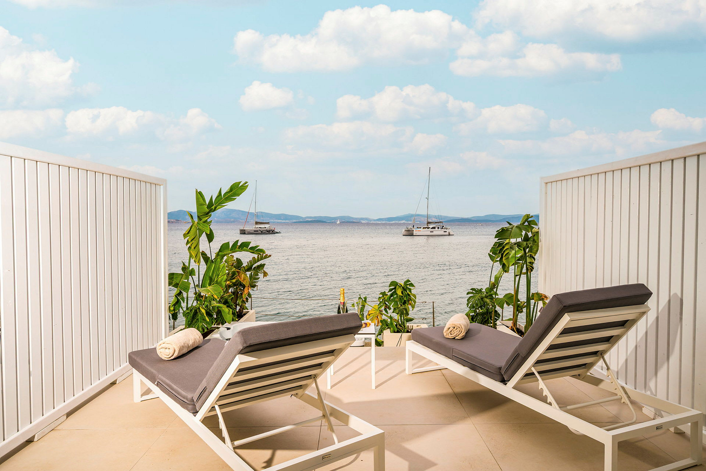 To solsenge med grå puder og håndklæder placeret på en terrasse med udsigt over havet og sejlbaåde. (AI-genereret tekst)
