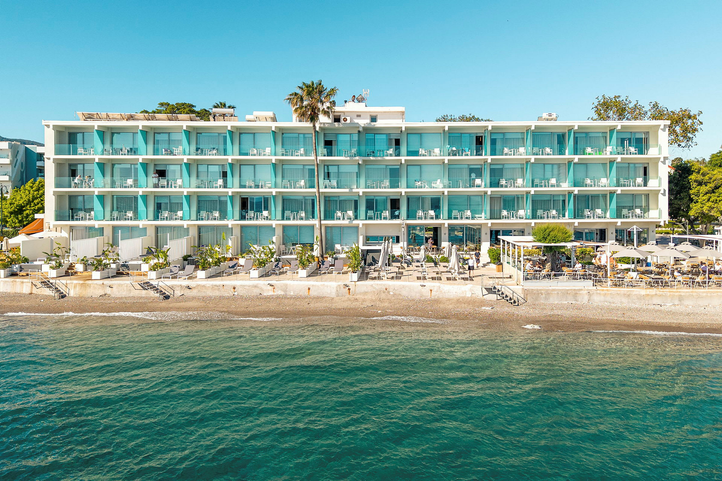 Et hotel ved stranden med en hvid facade og blå altaner. Der er en terrasse med parasoller og stole nedenfor, og havet ses i forgrunden. (AI-genereret tekst)
