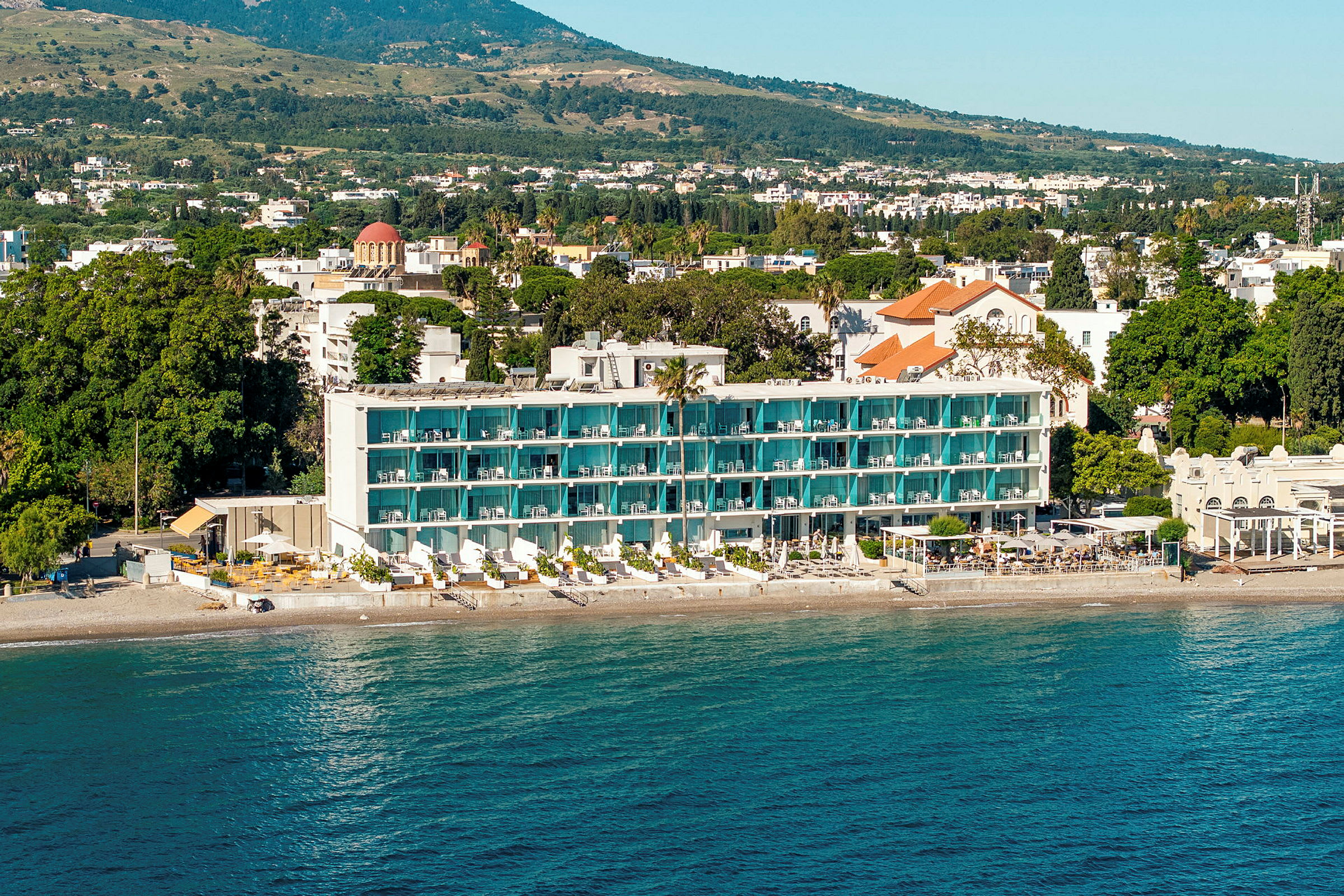 En hotelbygning med altaner og en strand foran, omgivet af grønne træer og et landskab i baggrunden. (AI-genereret tekst)