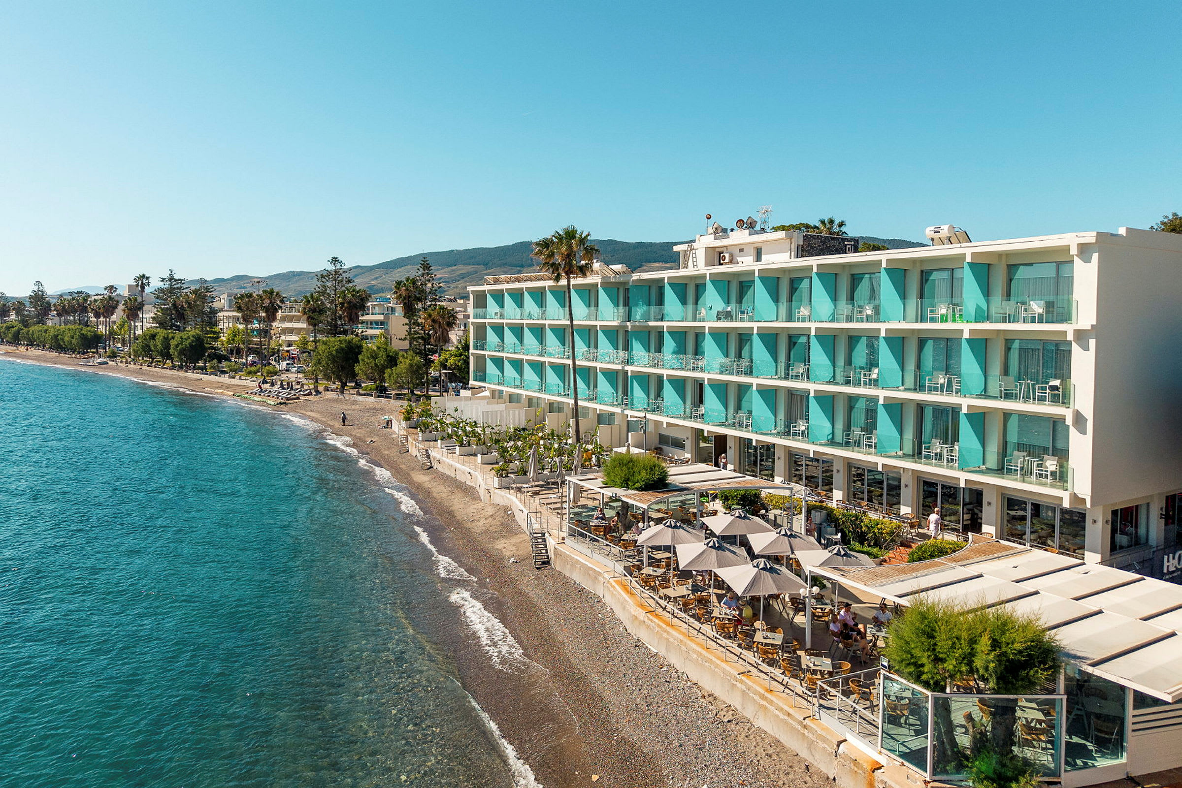 En strand med en moderne bygning med turkise altaner og en terrasse ud mod vandet. I forgrunden ses siddepladser med parasoller. (AI-genereret tekst)