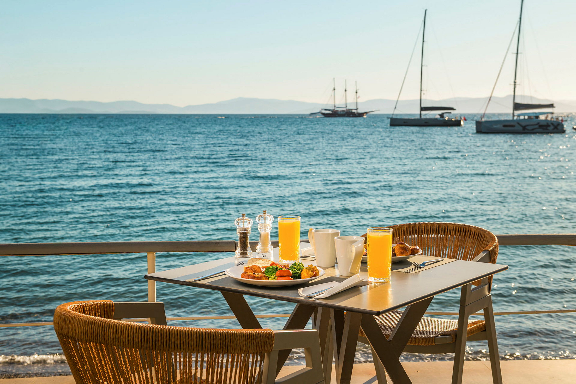 Et bord med morgenmad og juice står ved havet, med sejlbåde i baggrunden. (AI-genereret tekst)