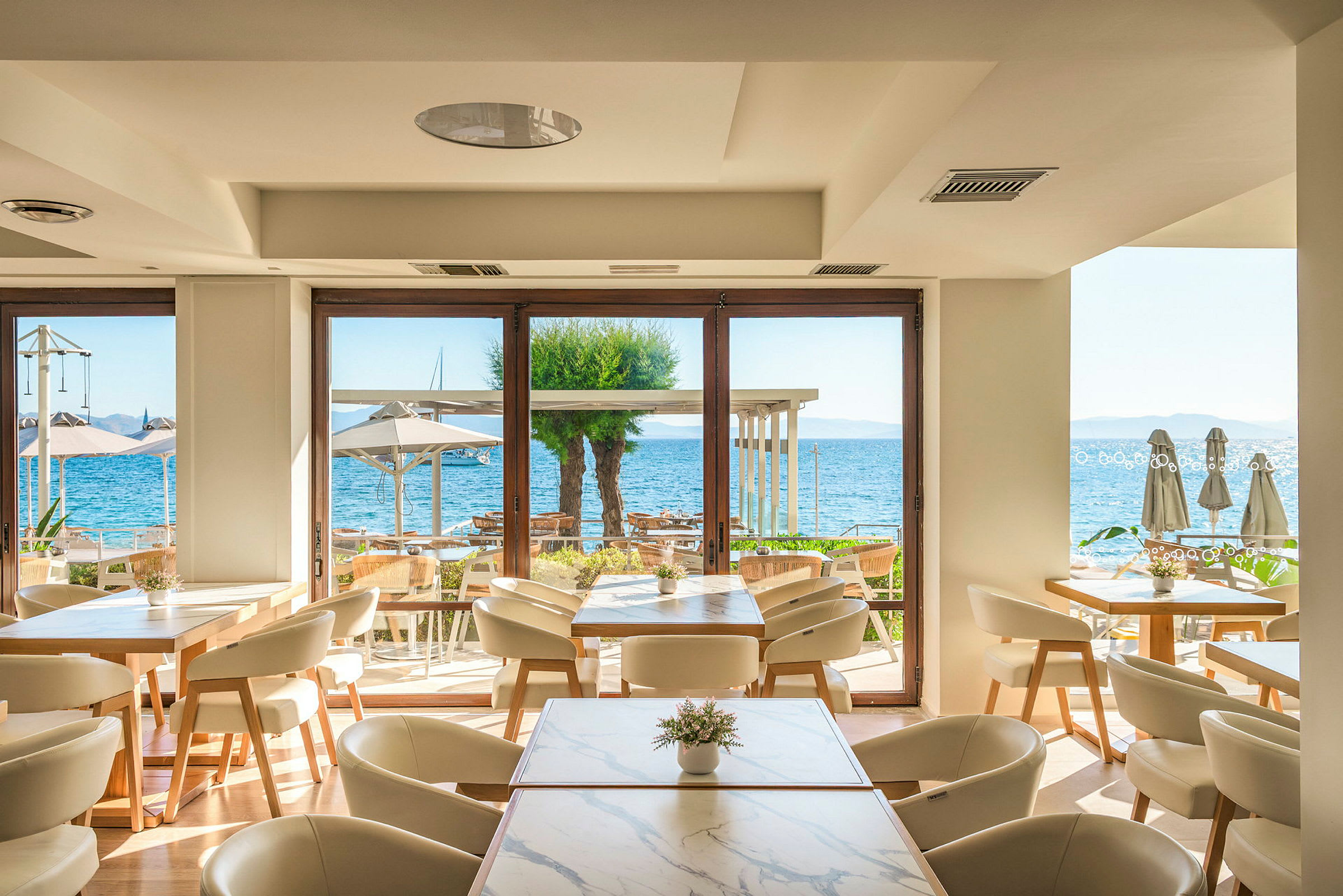 En lys og moderne restaurant med udsigt over havet gennem store vinduer. Inde står borde med stole, og udenfor ses parasoller og vandet. (AI-genereret tekst)