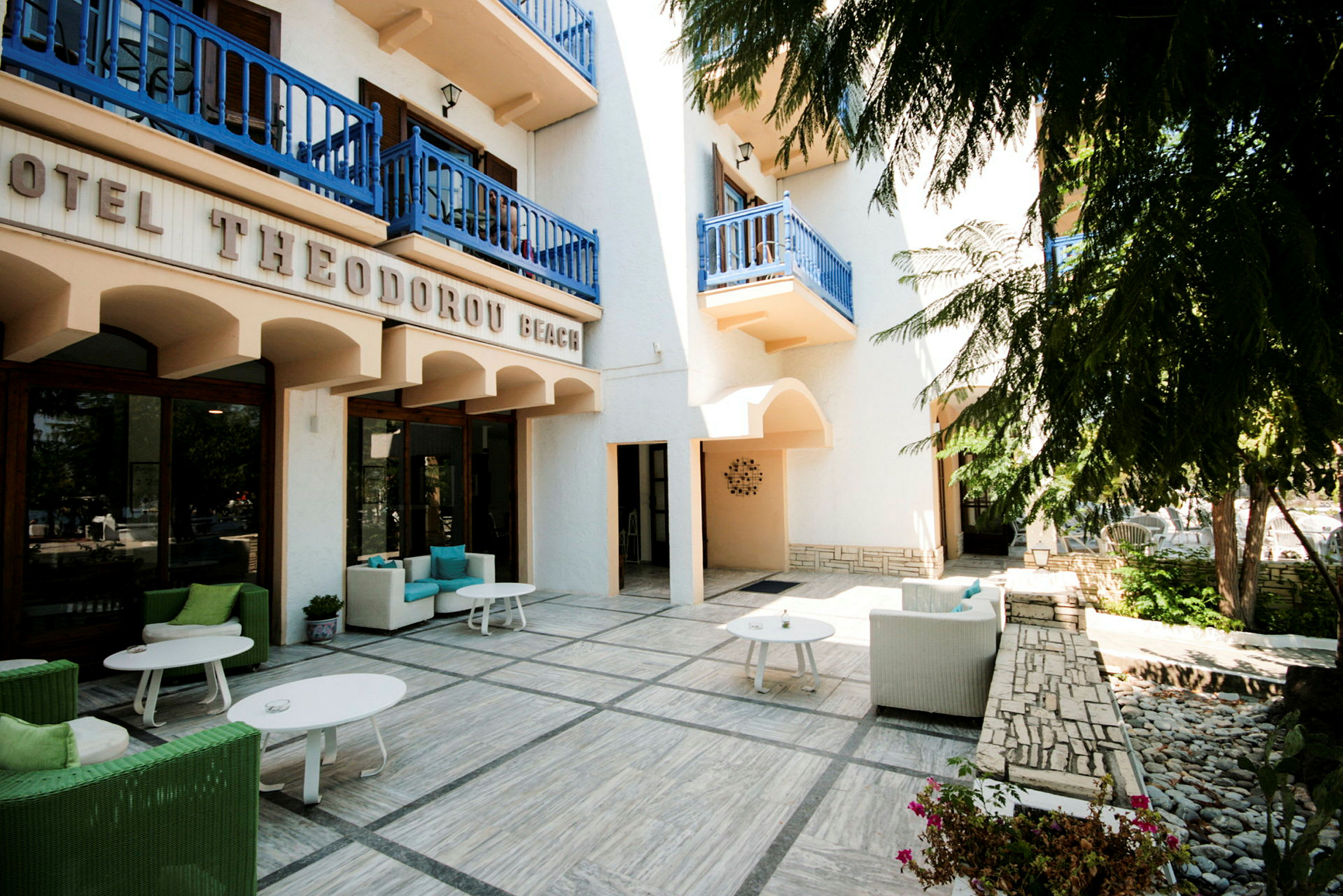 Udvendig af et hotel med terrasse med siddegrupper under blå altaner. (AI-genereret tekst)