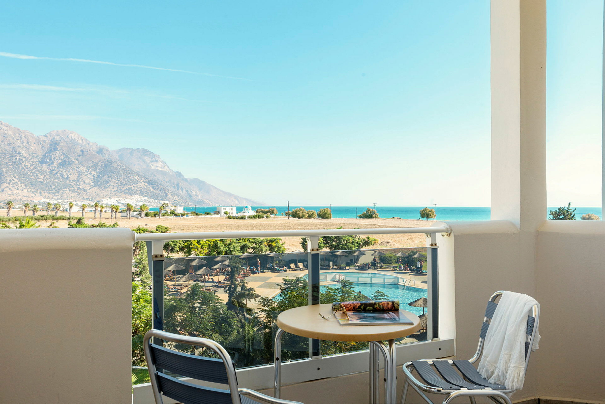 Udsigt fra en balkon med bord og stole mod en pool, landskab med bjerge og havet i baggrunden. (AI-genereret tekst)