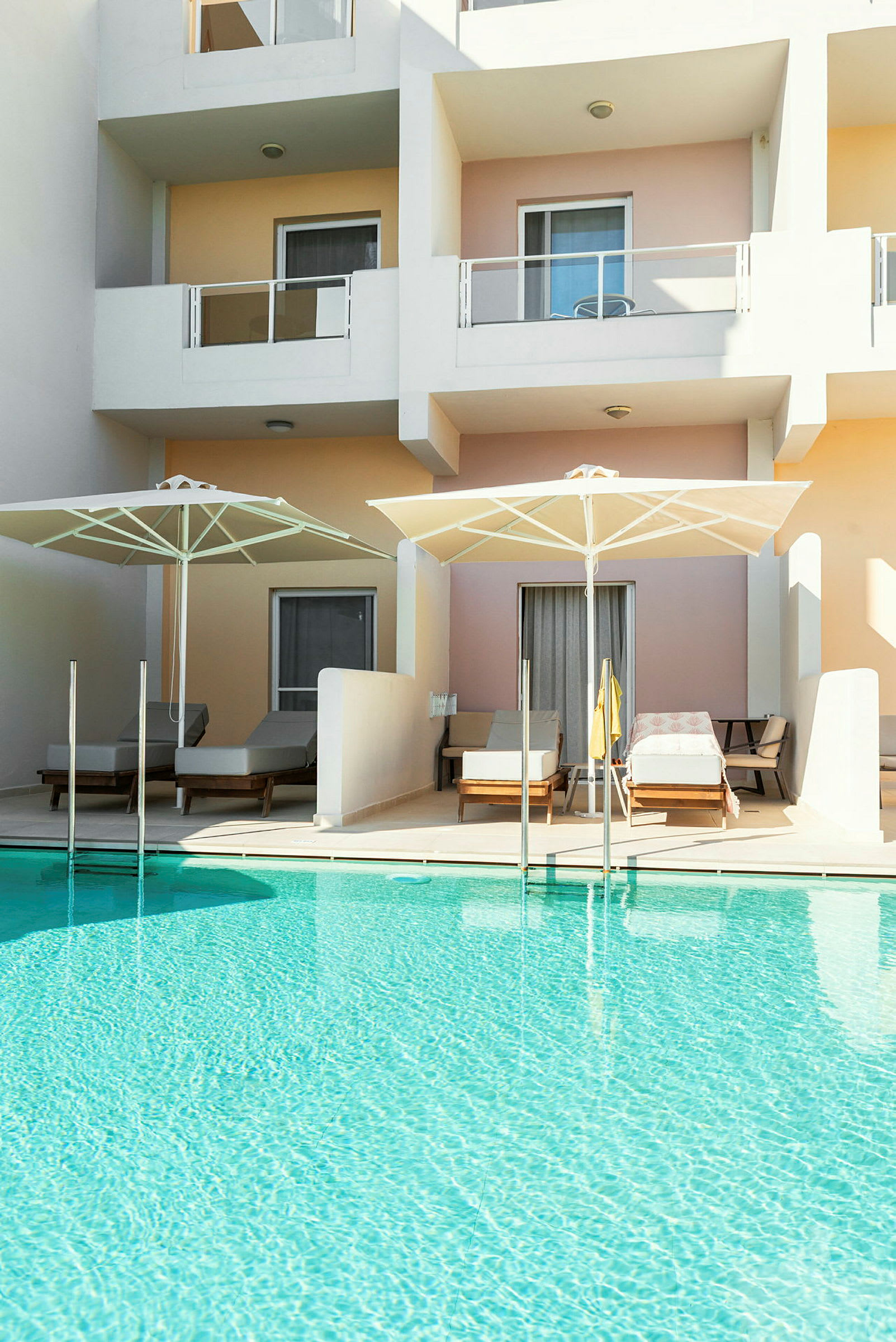 En swimmingpool foran lejlighedsbalkoner med liggestole og parasoller. (AI-genereret tekst)