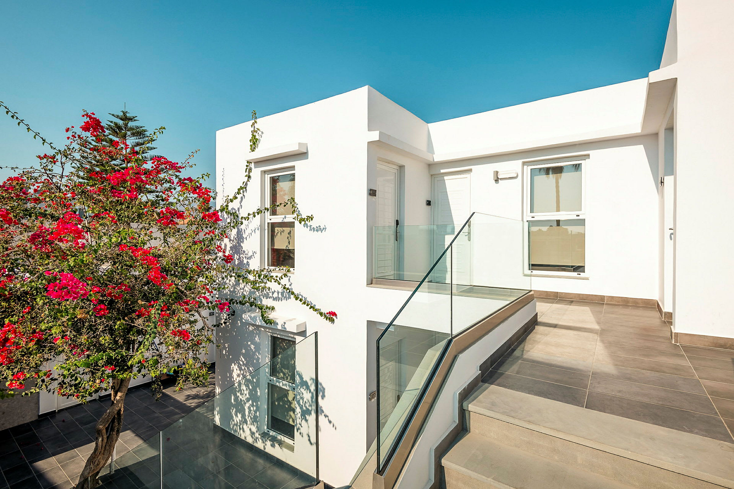 Moderne bygning med hvid facade, glasrækværk og blomstrende røde blomster. (AI-genereret tekst)