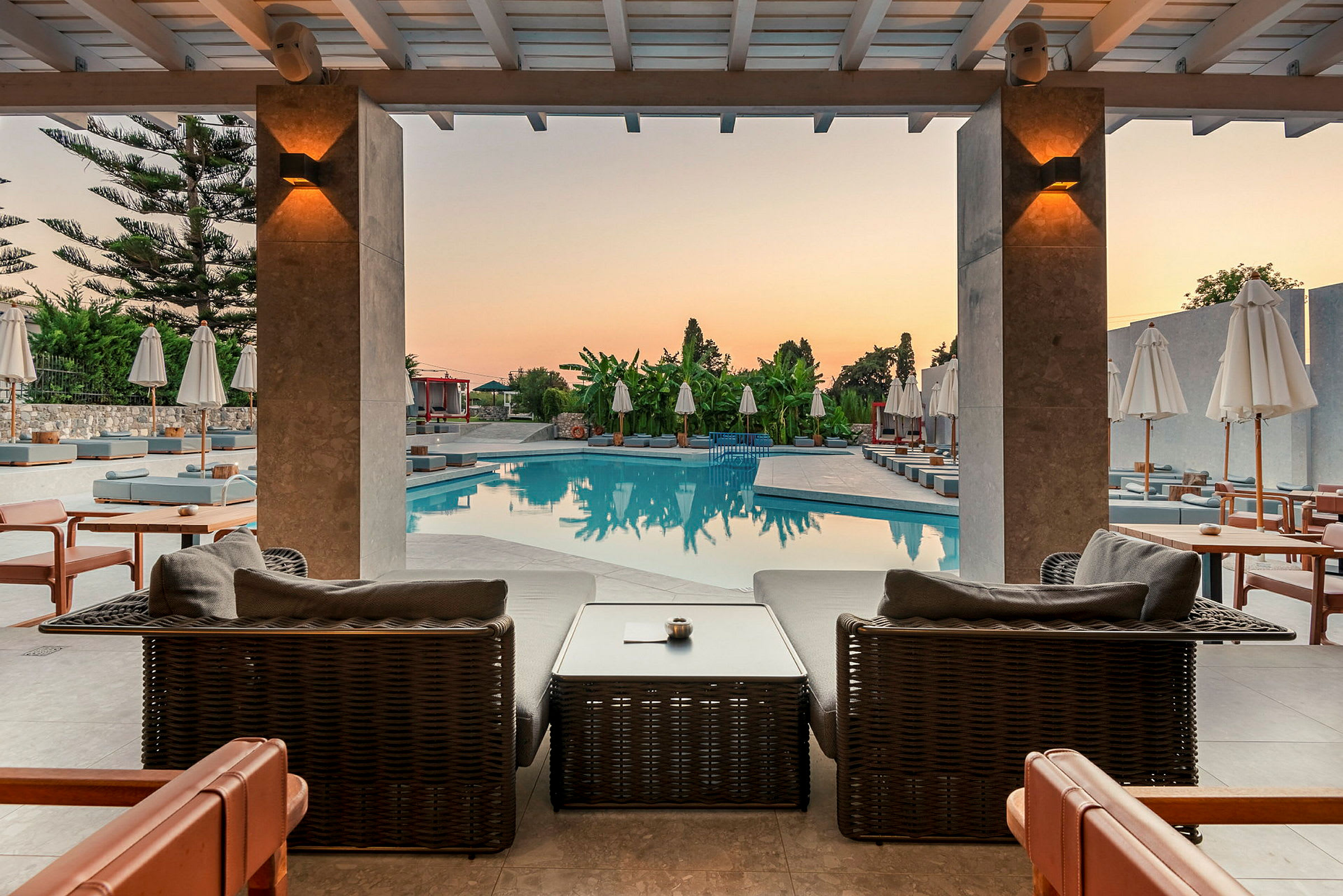 Solnedgang over en swimmingpool med liggestole og parasoller, set fra en terrasse med siddepladser. (AI-genereret tekst)