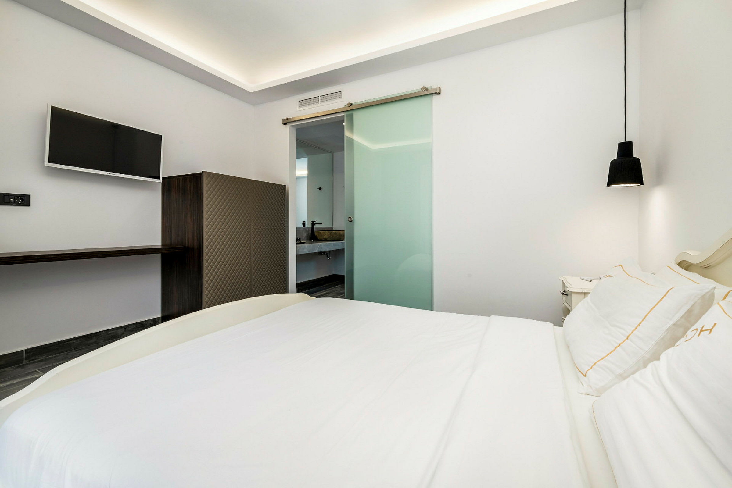 Et moderne hotelværelse med en dobbeltseng, et fladskærms-tv på væggen og et glasvindue ind til badeværelset. (AI-genereret tekst)