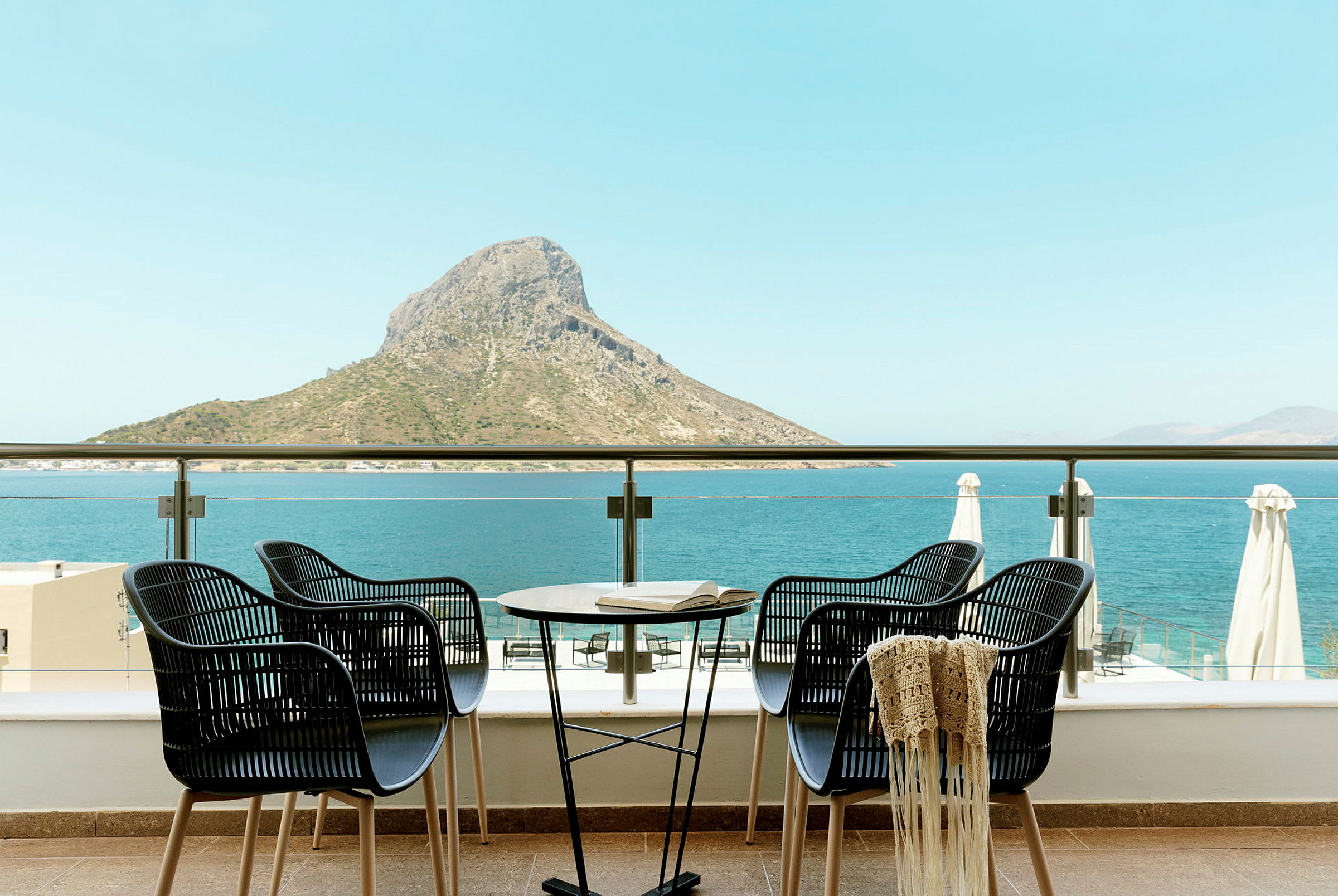 Fire stole og et bord på en terrasse med udsigt over havet og en bjergtop. (AI-genereret tekst)