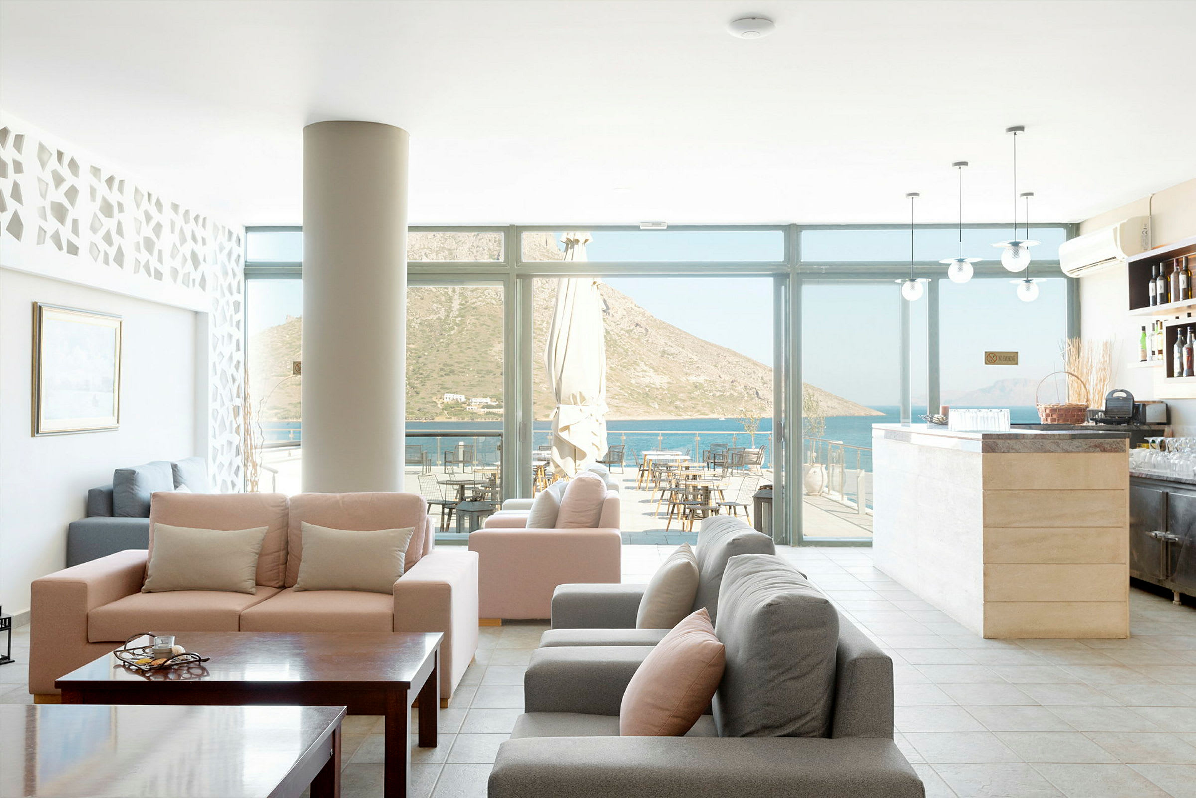 En hotellobby med komfortable sofaer, et bord og glasvægge, der viser udsigt over havet og bjergene. (AI-genereret tekst)