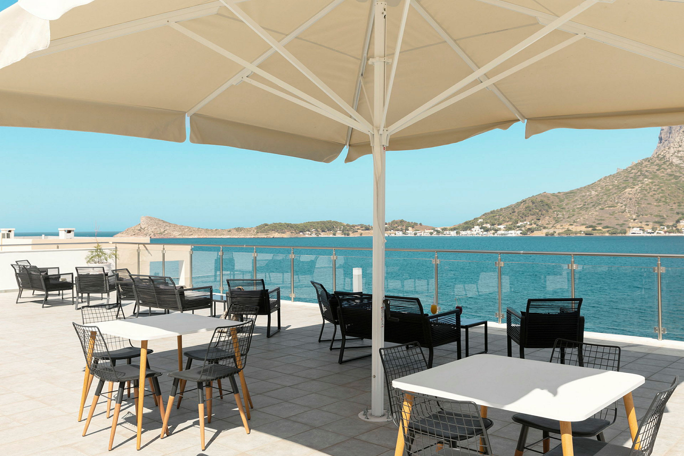 Udendørs serveringsområde med borde og stole under en stor parasol, med udsigt over havet og omkringliggende bakker. (AI-genereret tekst)