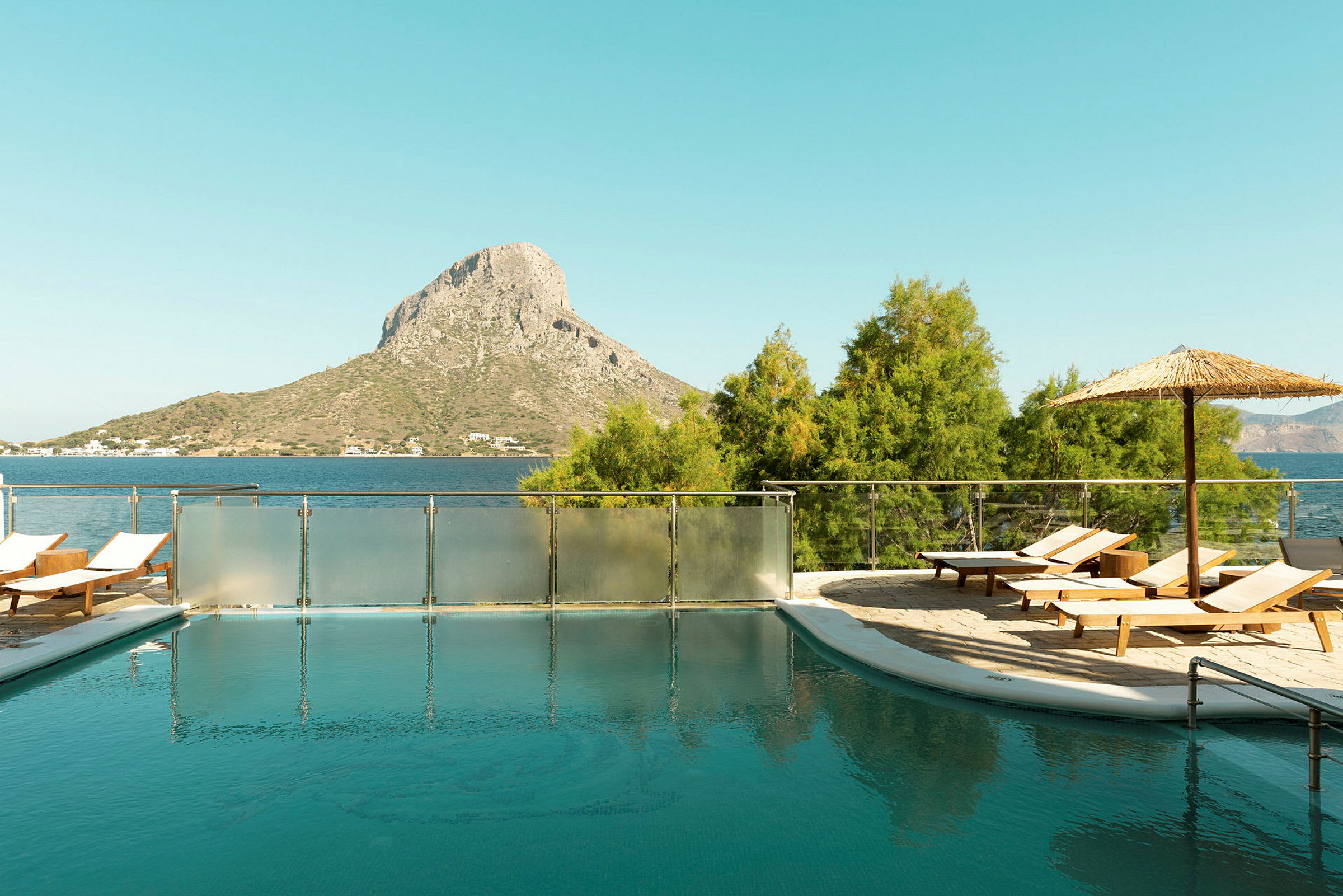 En udendørs pool med liggestole og parasol, beliggende tæt på havet med en bakkelignende høj i baggrunden. (AI-genereret tekst)