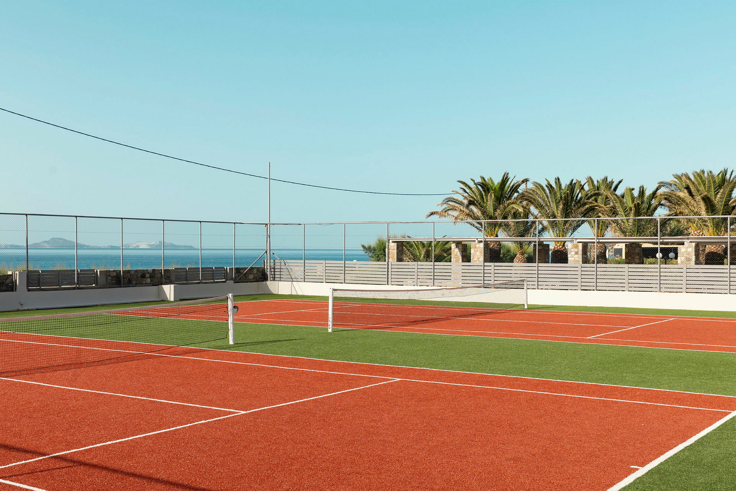 En udendørs tennisbane med rød belægning og grønt omgivelser. I baggrunden er der palmer og hav. (AI-genereret tekst)