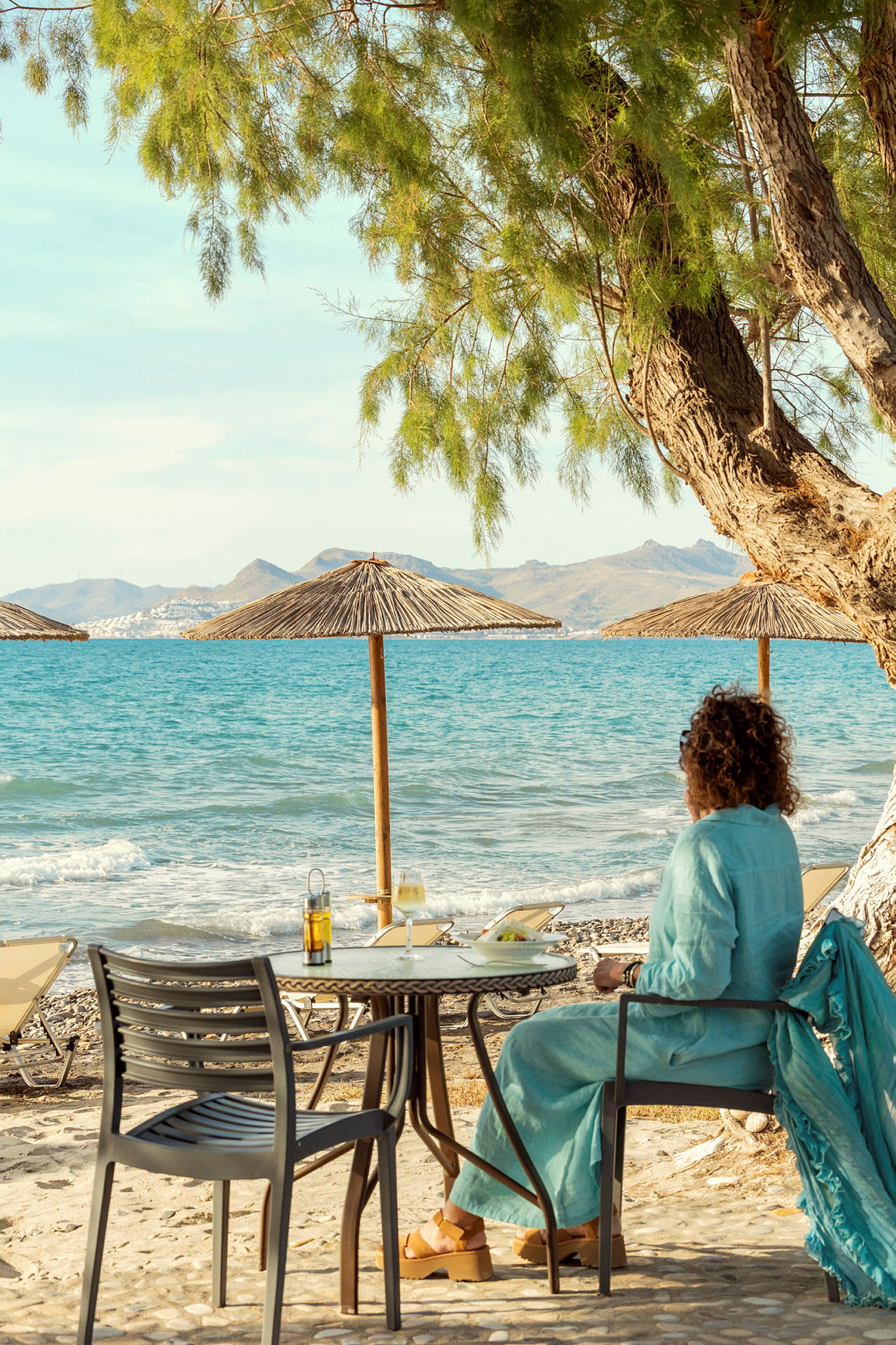 Person sidder ved et bord på en stenstrand med udsigt over havet og bjerge i baggrunden. (AI-genereret tekst)