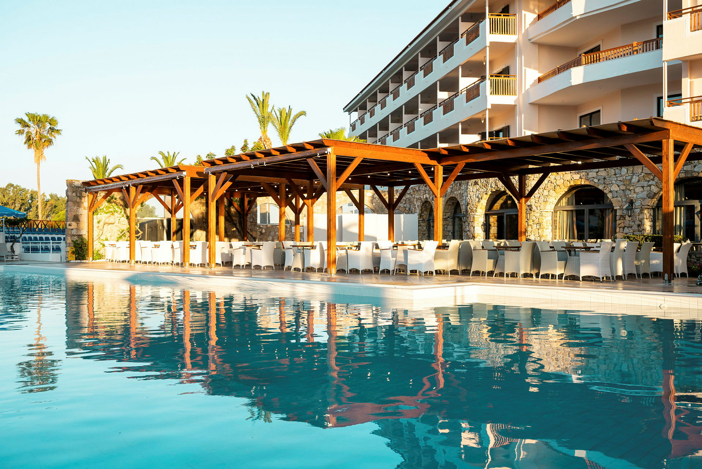 En udendørs swimmingpool med spejlinger af en bygning og siddepladser under en træpergola. (AI-genereret tekst)