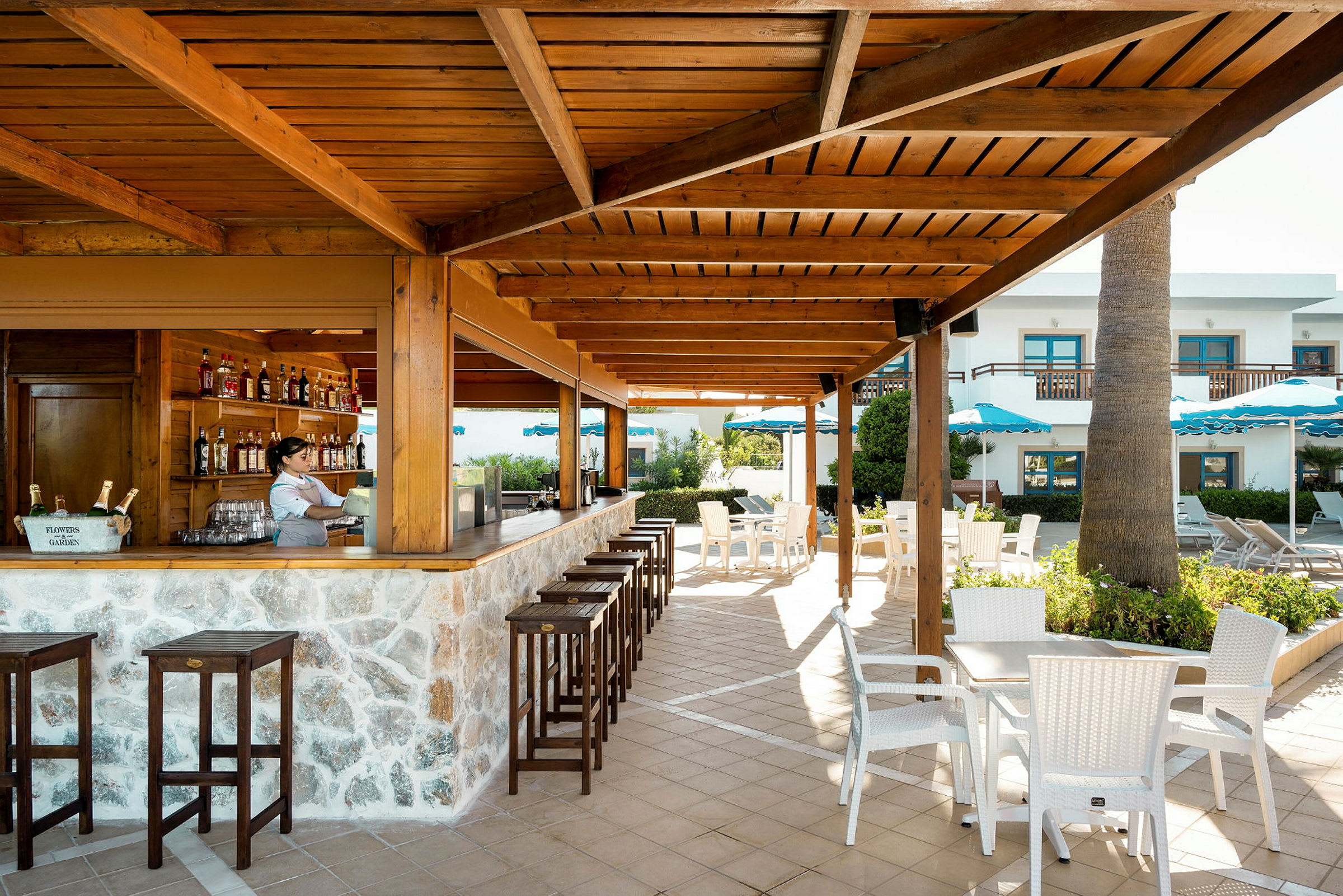 Billedet viser et udendørsområde med en bar under et trætag, stole og borde i et terrasseomgivelser. (AI-genereret tekst)