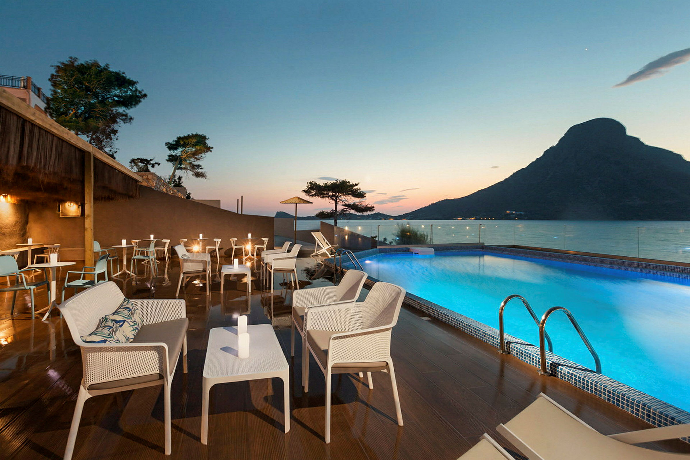 Udendørs terrasse med siddepladser og borde ved en oplyst pool med udsigt over havet i skumringen. (AI-genereret tekst)