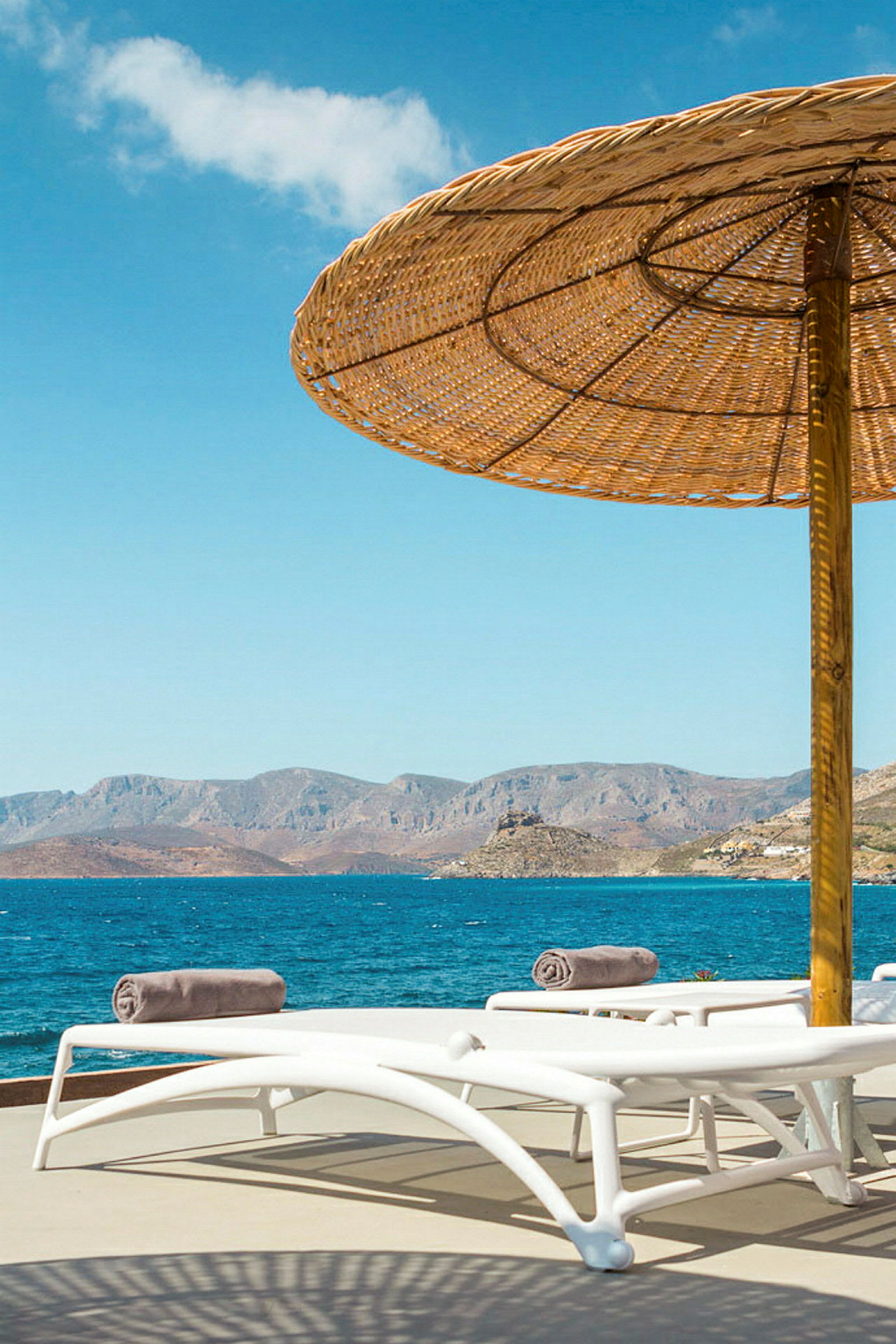 Solstole under en parasol ved havet med bjerge i baggrunden. (AI-genereret tekst)