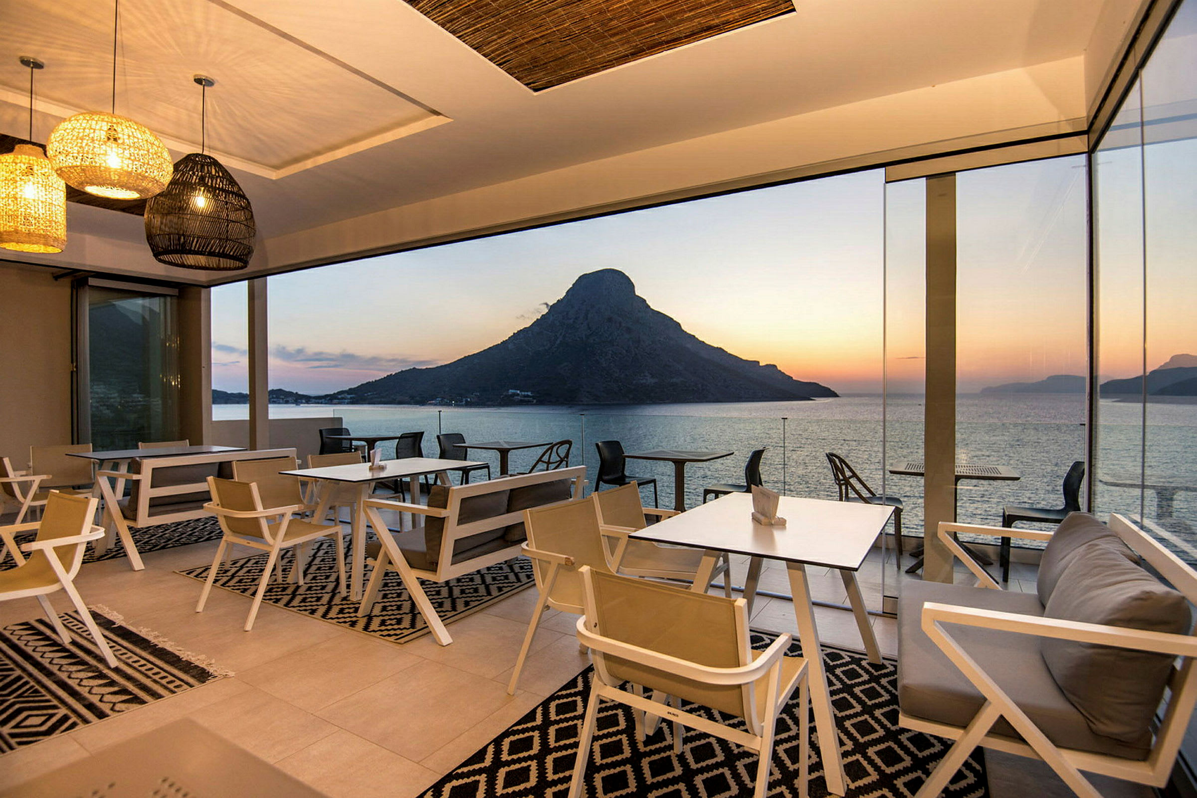 Restaurant med glasvægge og udsigt over havet og bjerge ved solnedgang. (AI-genereret tekst)