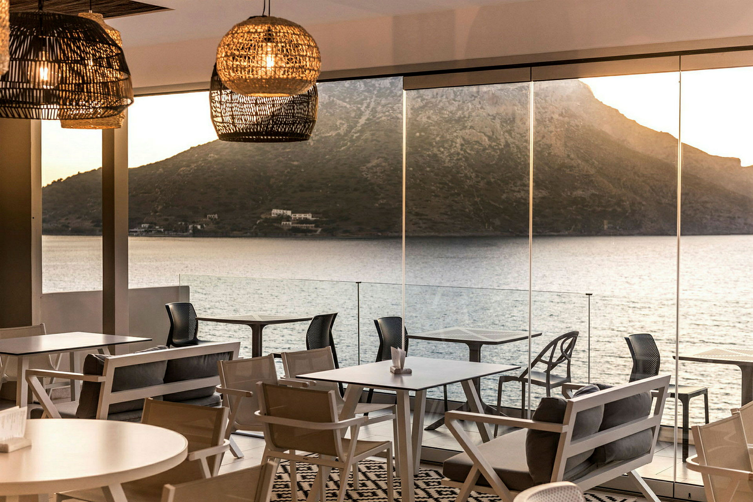 Udsigt over hav og bjerge i baggrunden fra en restaurant med store glasvinduer og moderne møbler. (AI-genereret tekst)