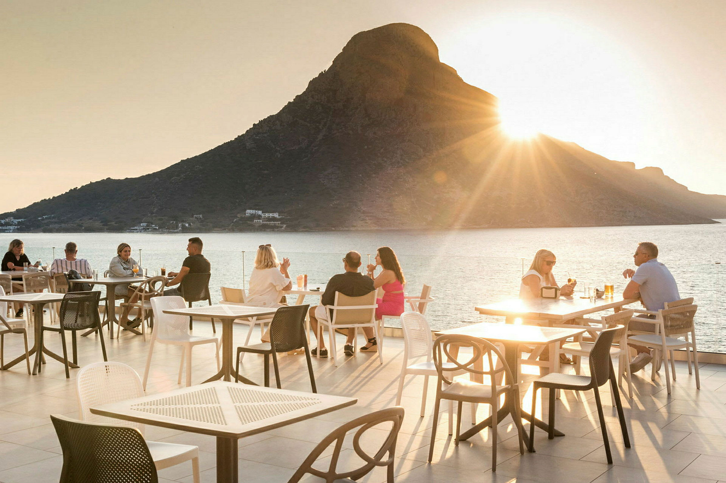En udendørs terrasse med siddepladser og et bjerg ved havet i baggrunden ved solnedgang. (AI-genereret tekst)