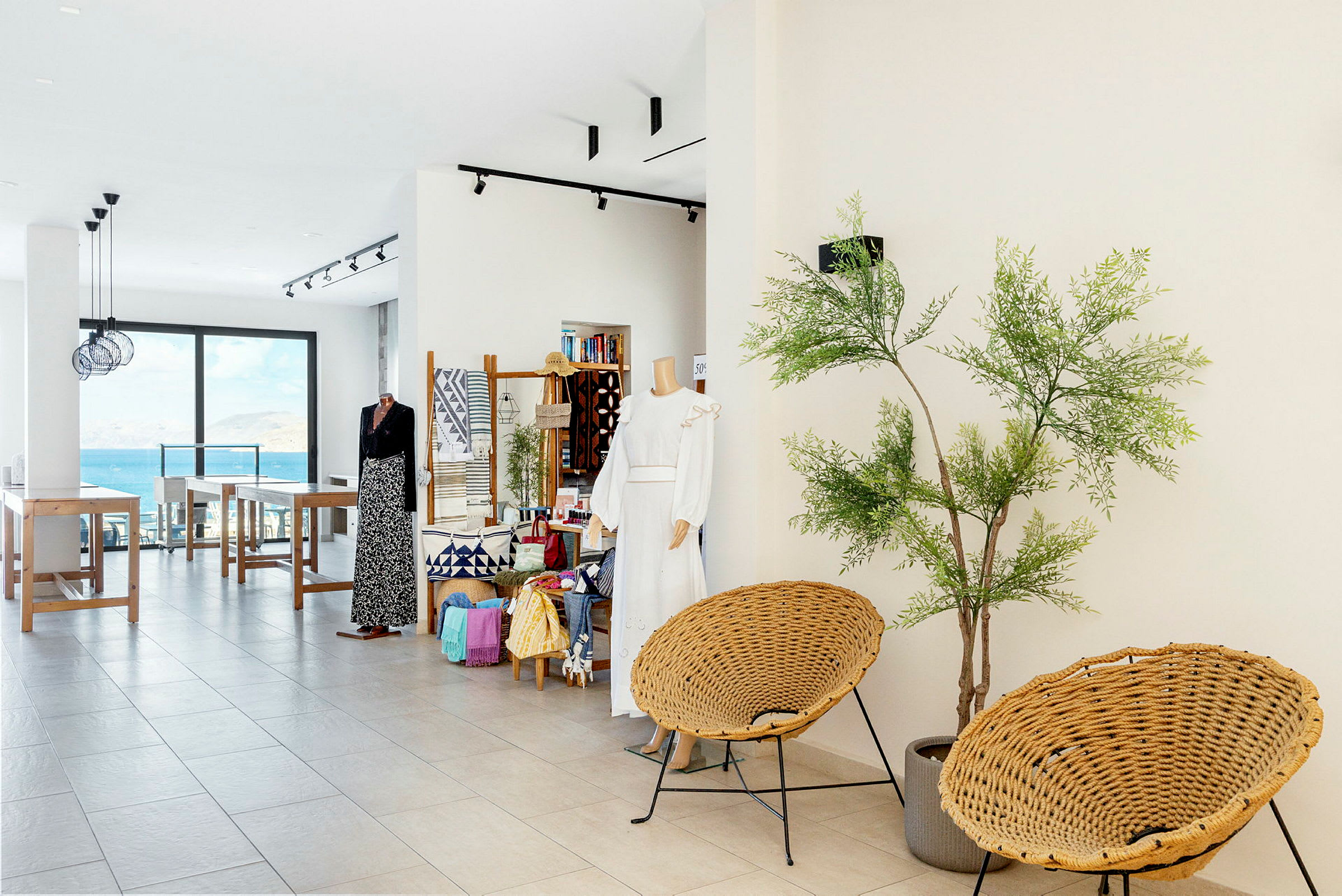 Et butikslignende område med tøj, tilbehør og dekorationer samt siddepladser og udsigt over havet. Planter bruges til at skabe en hyggelig atmosfære. (AI-genereret tekst)