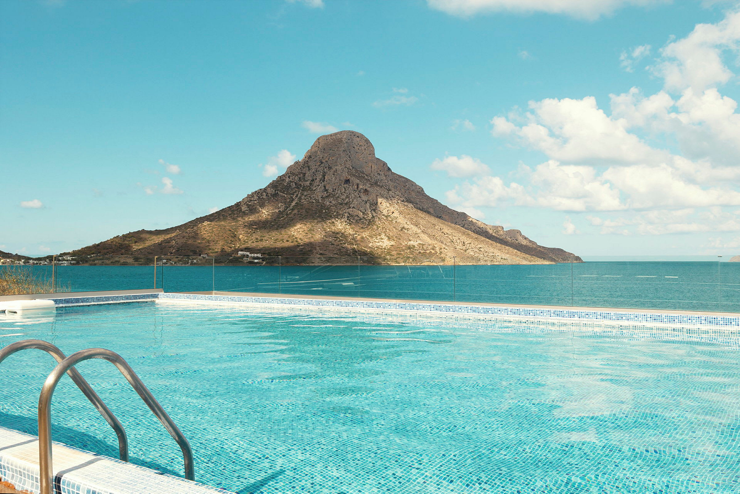 En pool med klart blåt vand foran et bjerg ved havet under en klar himmel. (AI-genereret tekst)