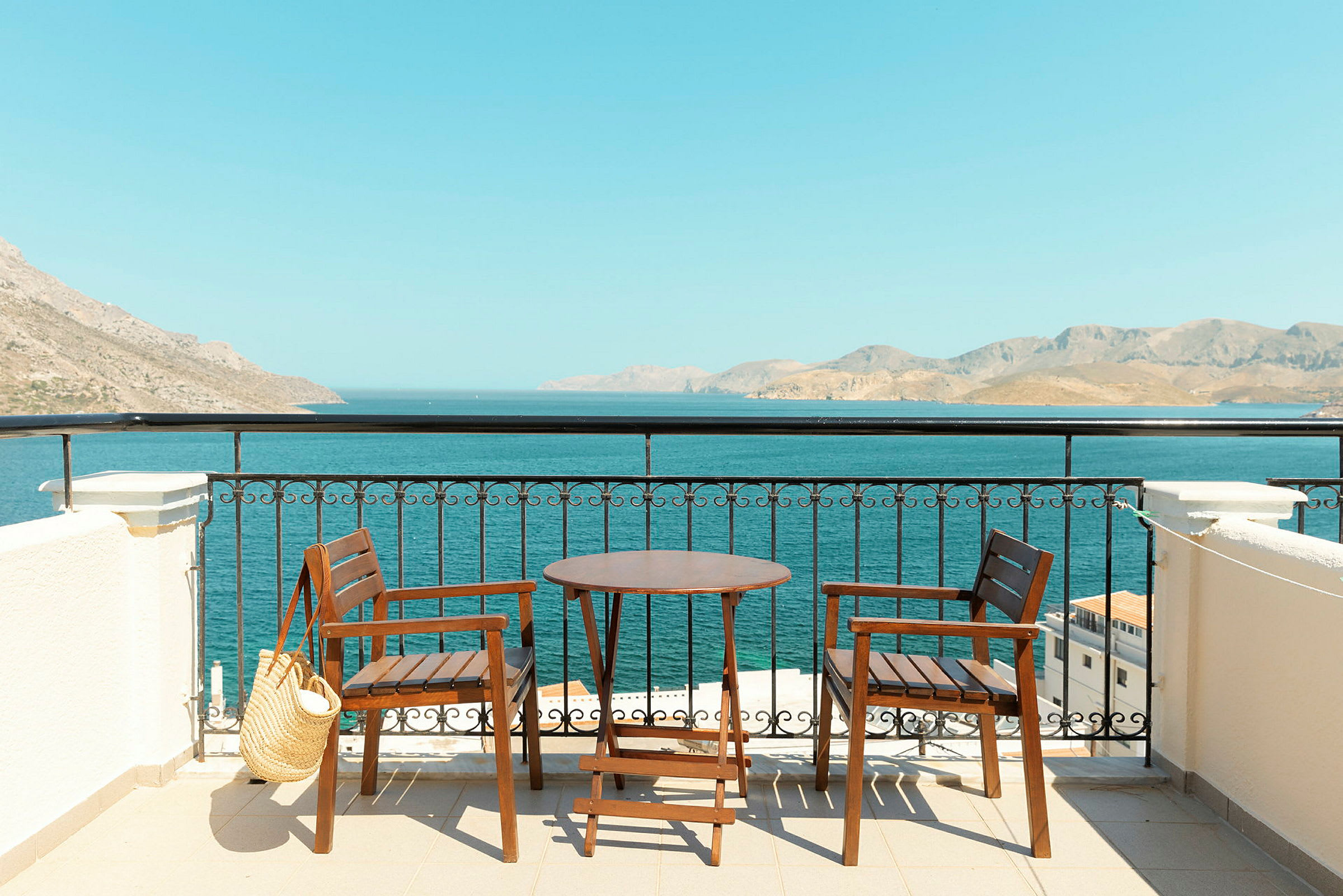 To træstole og et bord på en terrasse med udsigt til havet og bjerge i det fjerne. (AI-genereret tekst)