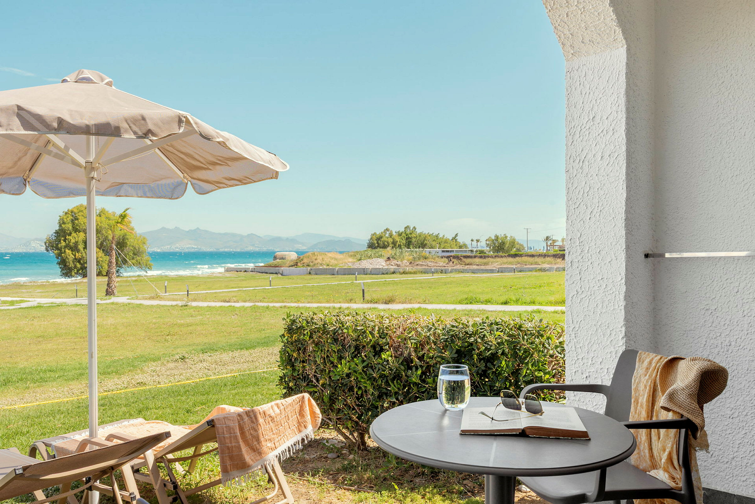Udsigt fra terrasse med bord, stole, parasol og grønt område mod havet. (AI-genereret tekst)