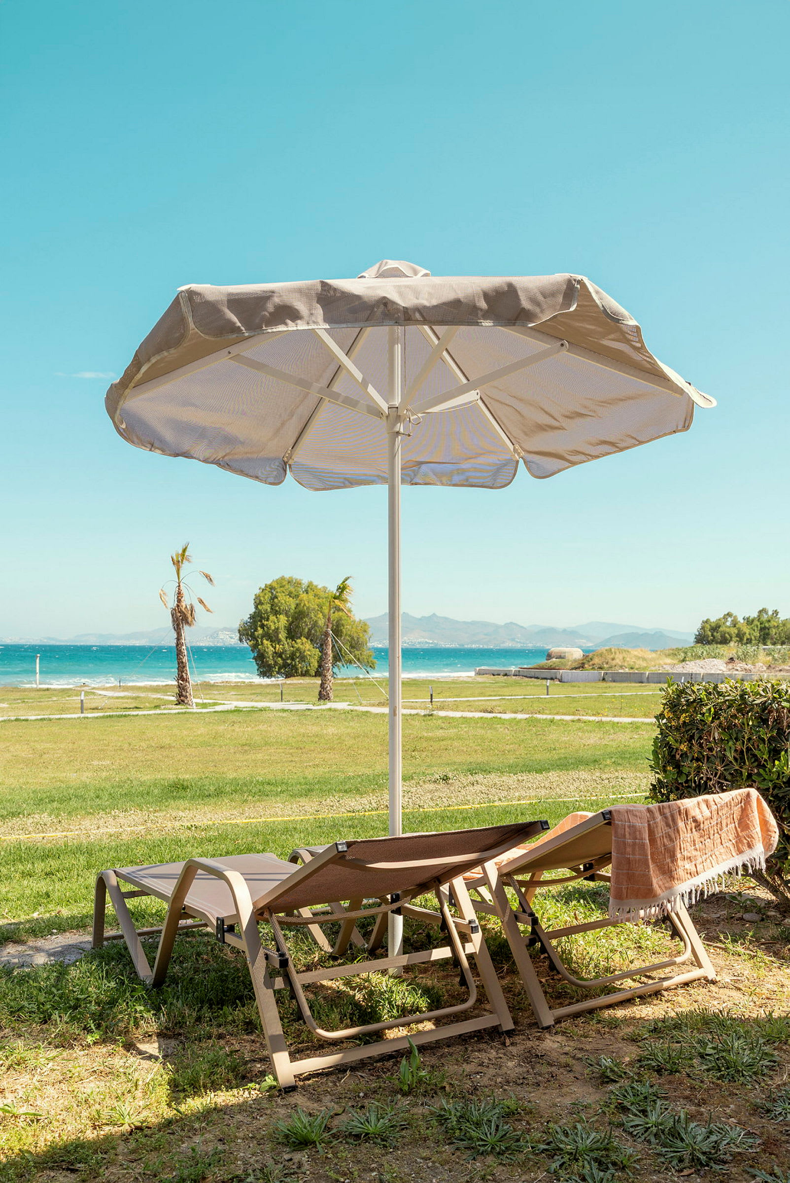 To liggestole står under en parasol med udsigt til havet og grønne områder. (AI-genereret tekst)