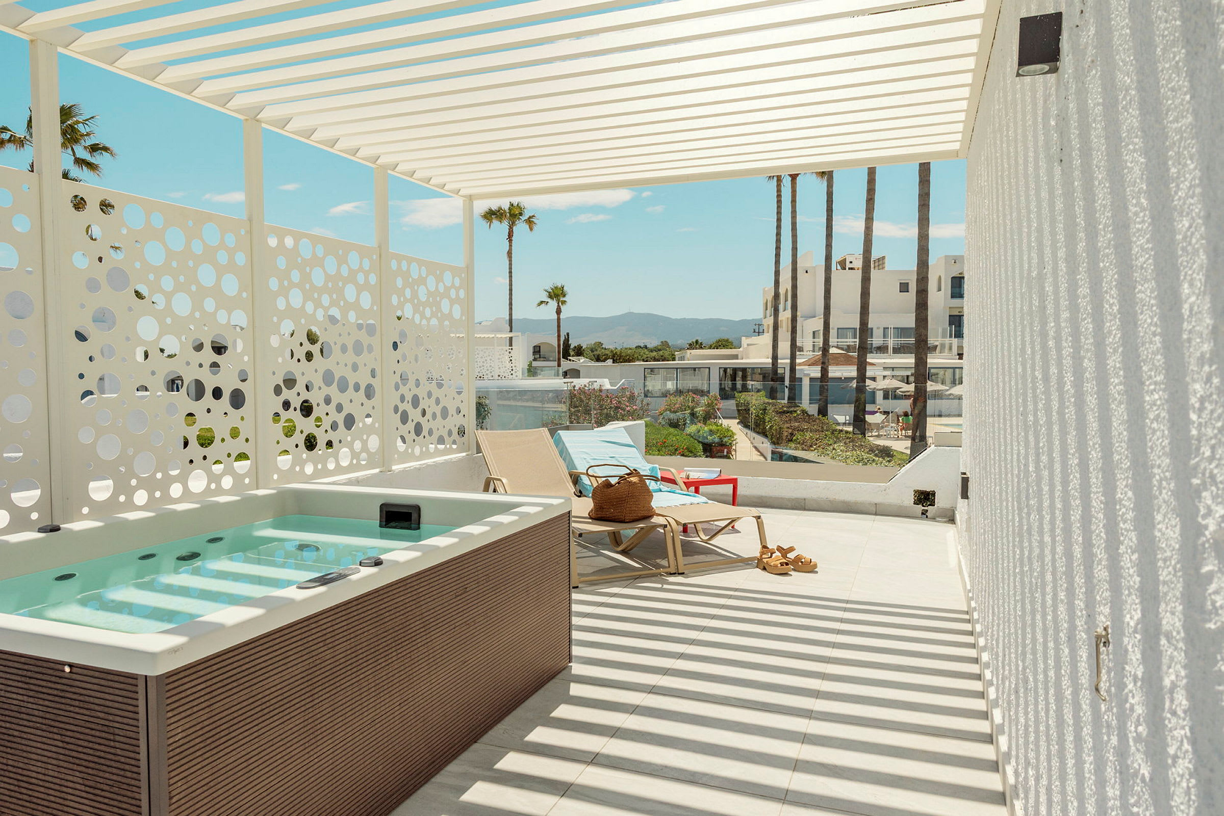 En terrasse med jacuzzi, liggestole og udsigt til palmer og bygninger i baggrunden. (AI-genereret tekst)