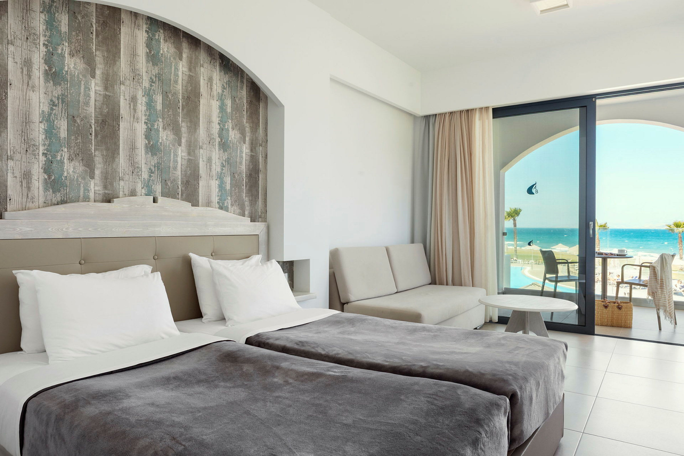 Hotelværelse med dobbeltsenge, et siddeområde og udsigt over havet gennem en glasdør. (AI-genereret tekst)