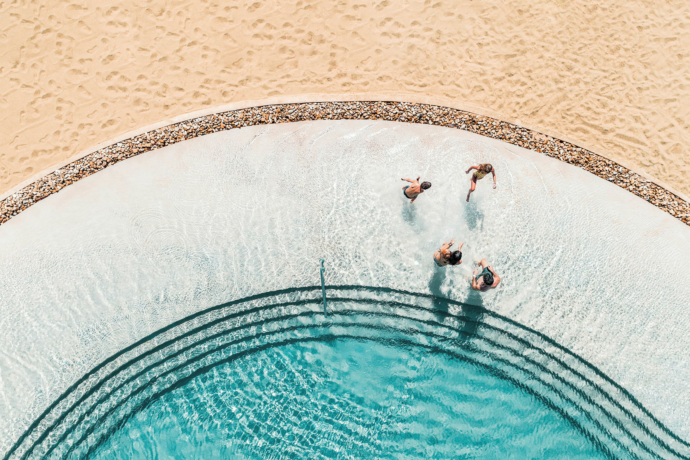 Udsigt ovenfra af en swimmingpool med mennesker, der svømmer nær en strand med sand. (AI-genereret tekst)