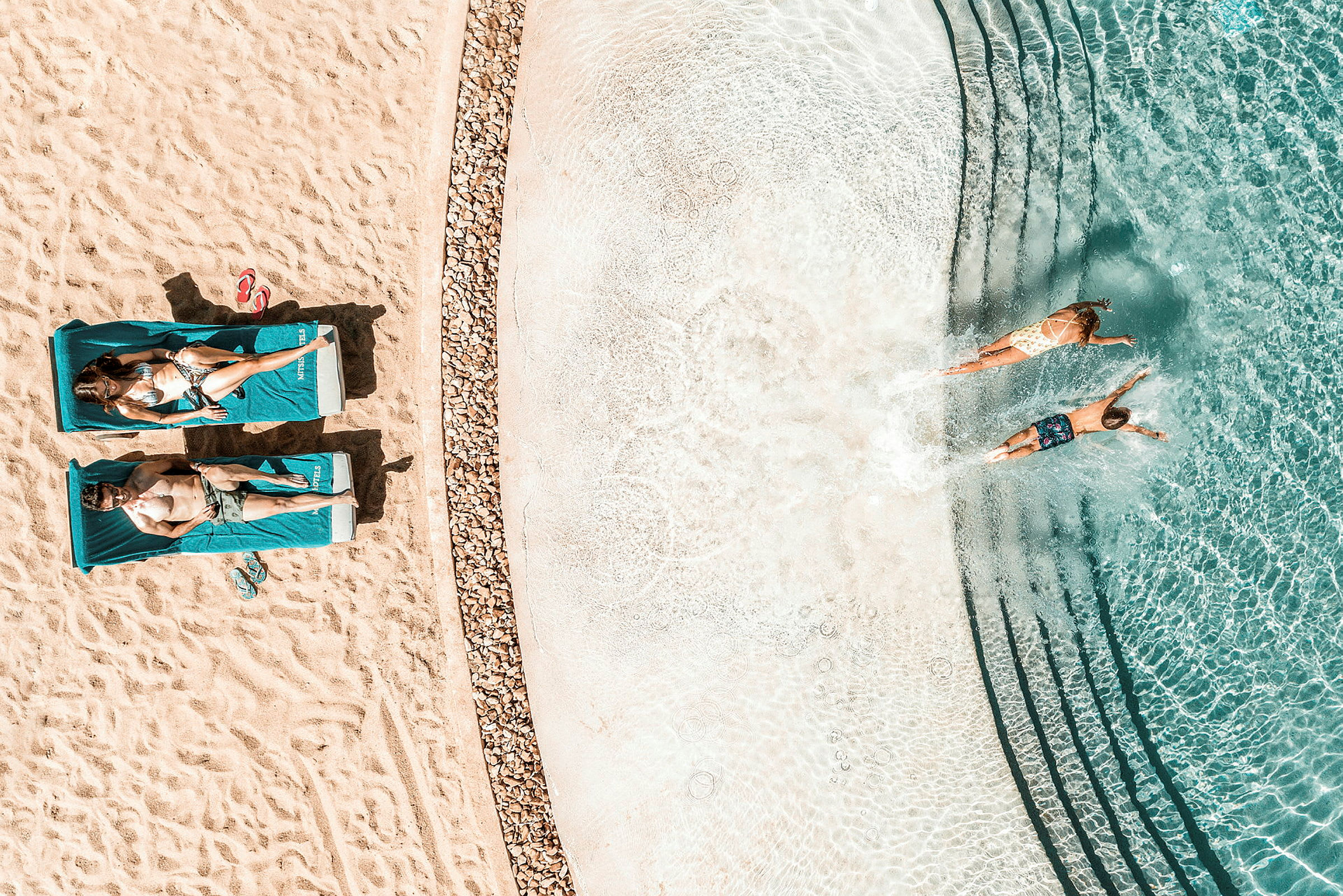 Luftfoto af to personer, der ligger på liggestole på en strand ved siden af en klarblå pool, hvor to andre personer svømmer. (AI-genereret tekst)