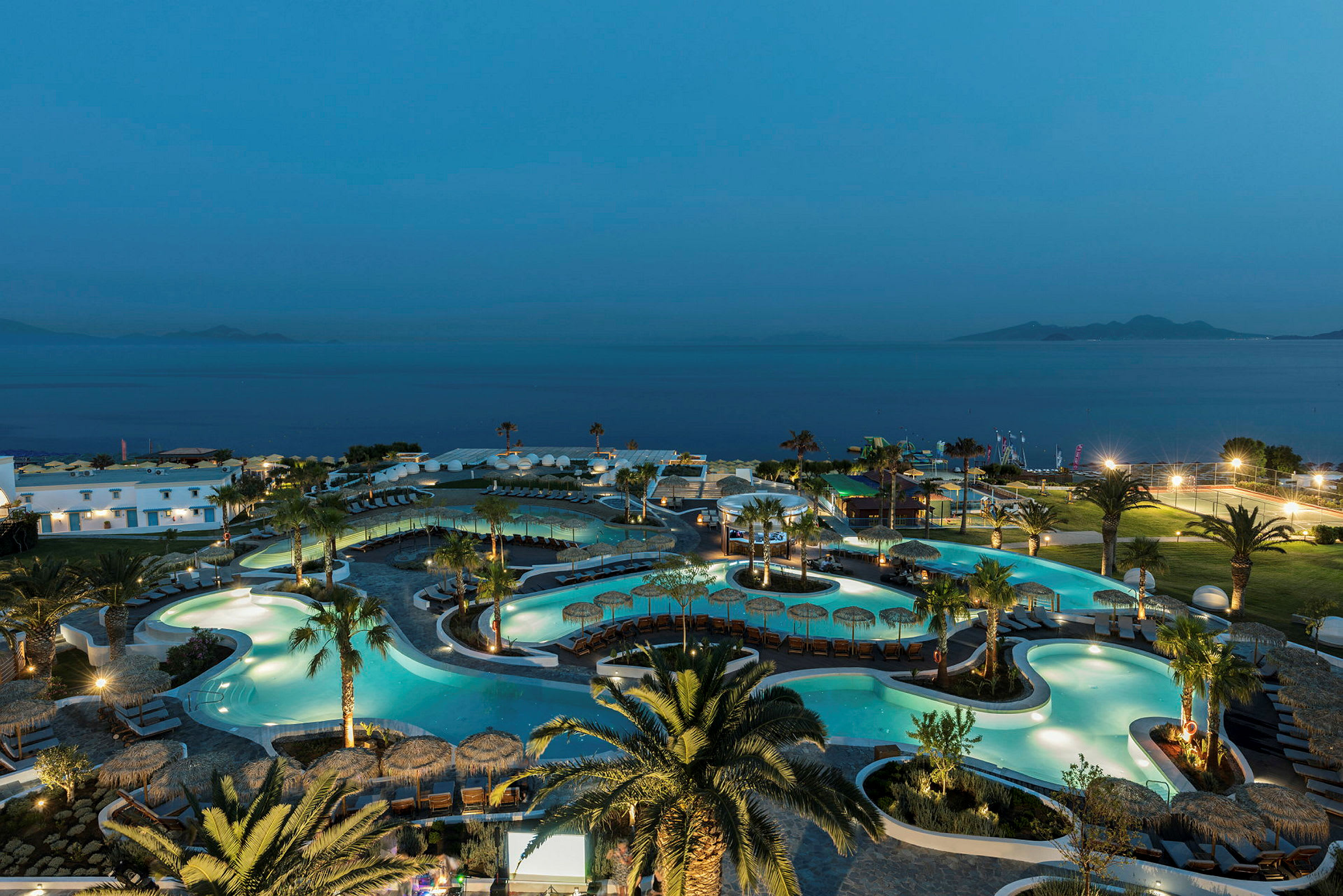 Udsigt over et oplyst hotelområde med pools og palmer om aftenen, med havet i baggrunden. (AI-genereret tekst)