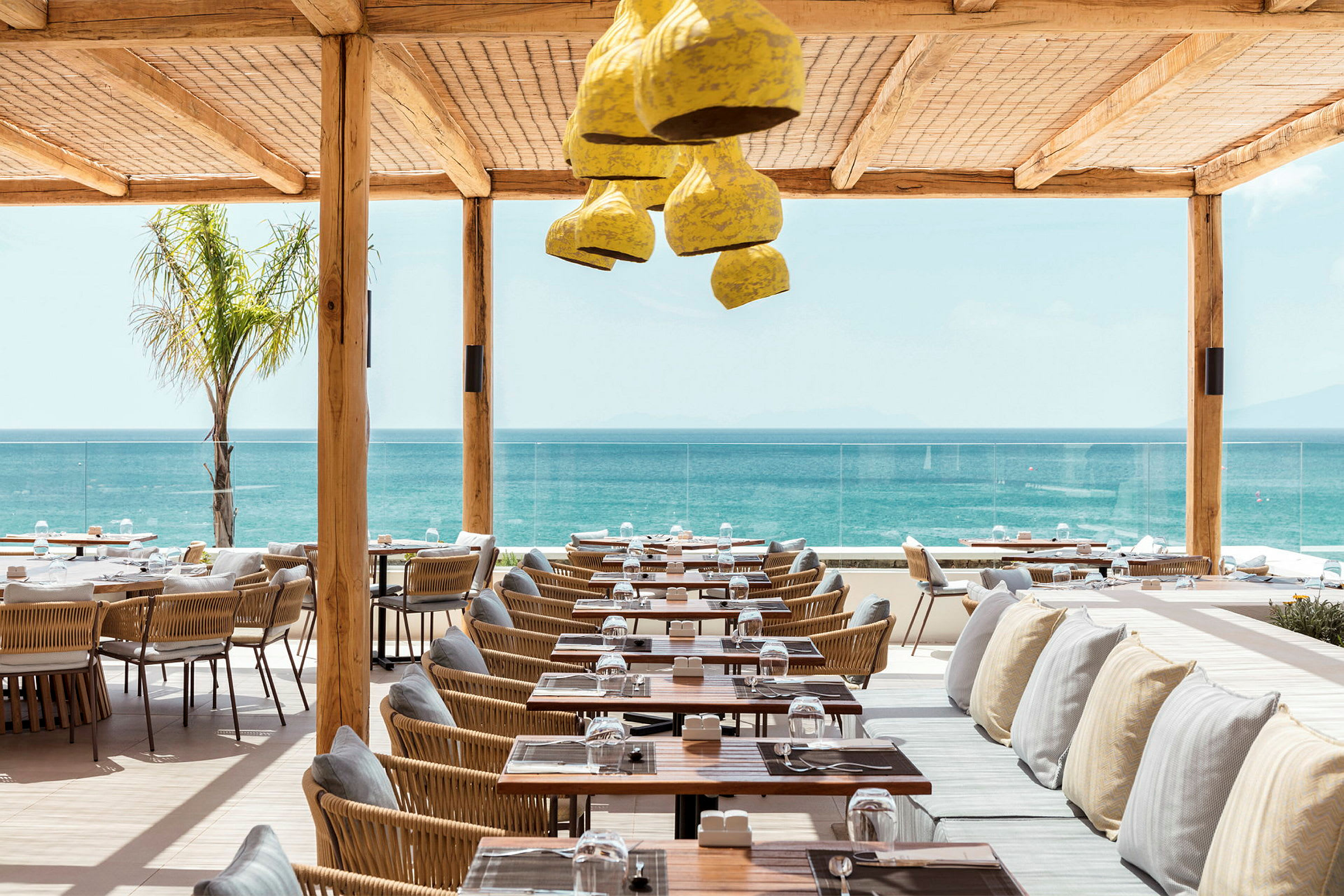 Restaurant med havudsigt, med borde og stole i træ og flet samt bænke med puder under et beskyttende tag. (AI-genereret tekst)