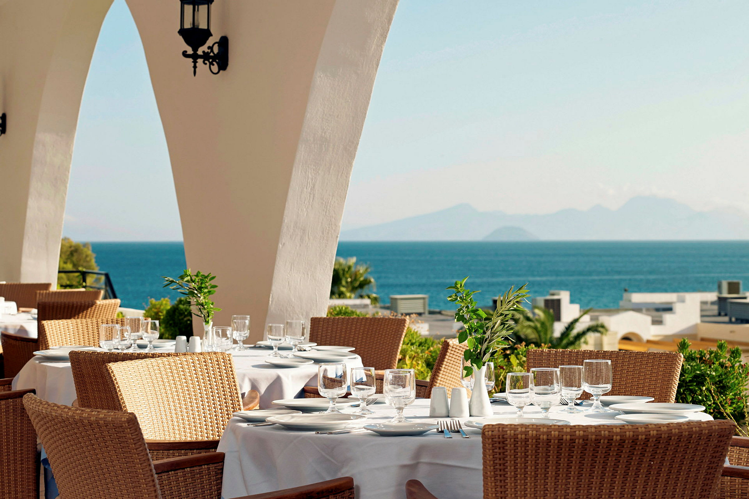 En restaurant med borde dækket med hvide duge, en solrig udsigt over havet og bjerge i baggrunden. (AI-genereret tekst)
