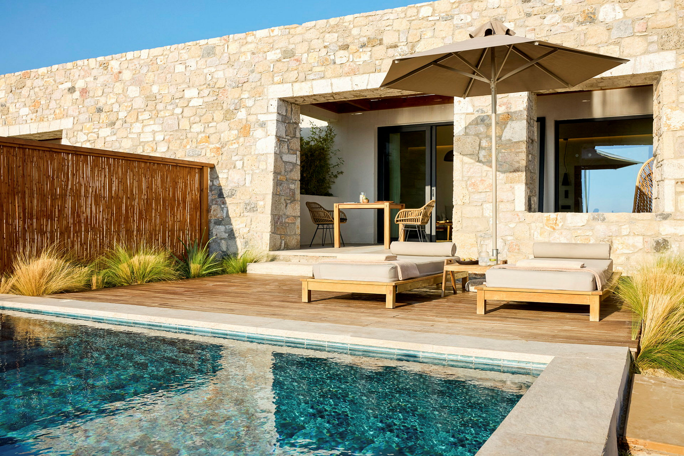 En udvendig af en stenhusbygning med terrasse, liggestole, parasol og pool. (AI-genereret tekst)